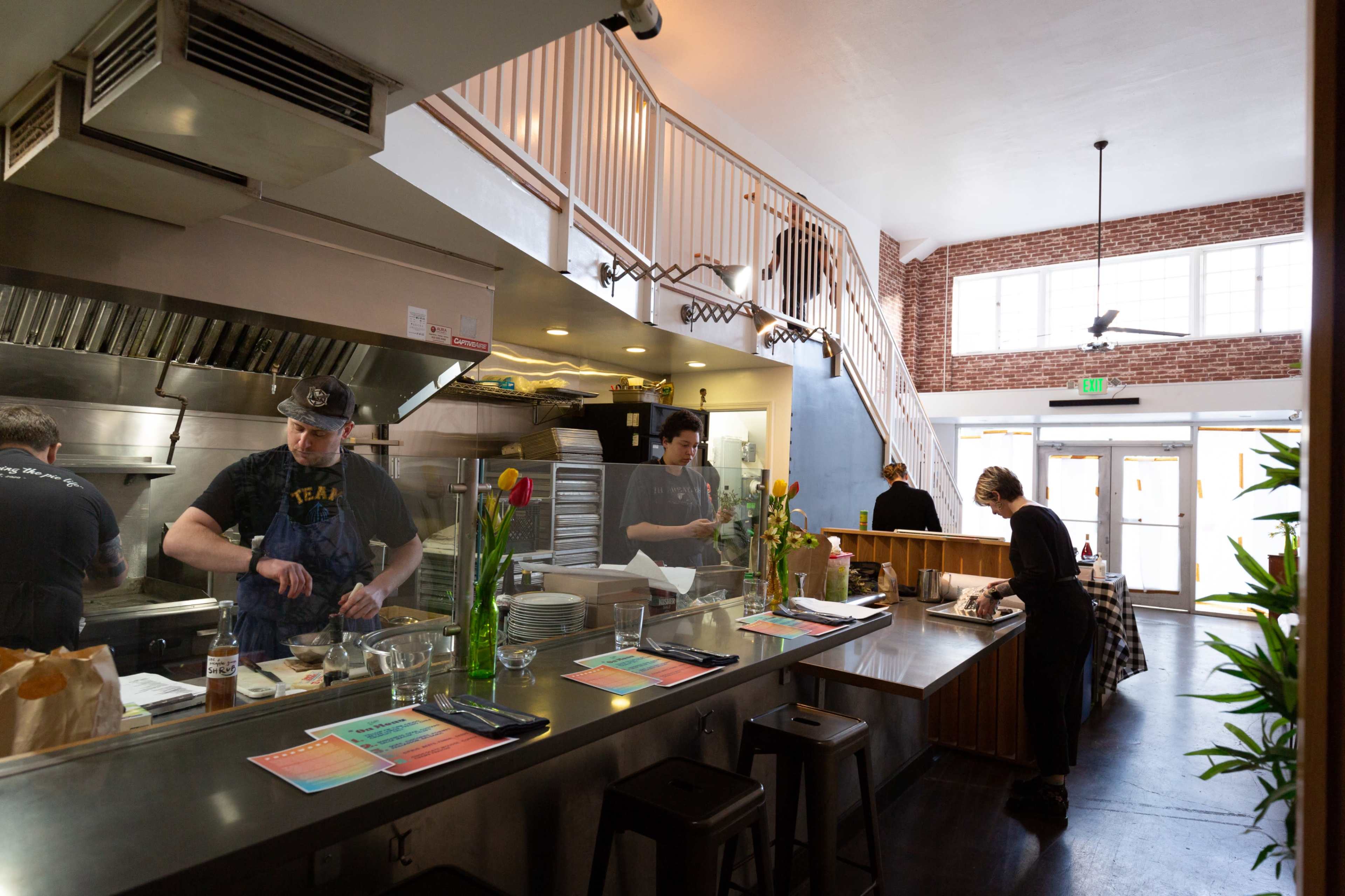A busy restaurant kitchen with chefs preparing food and staff working at a counter, while a staircase leads to an upper level in the background.