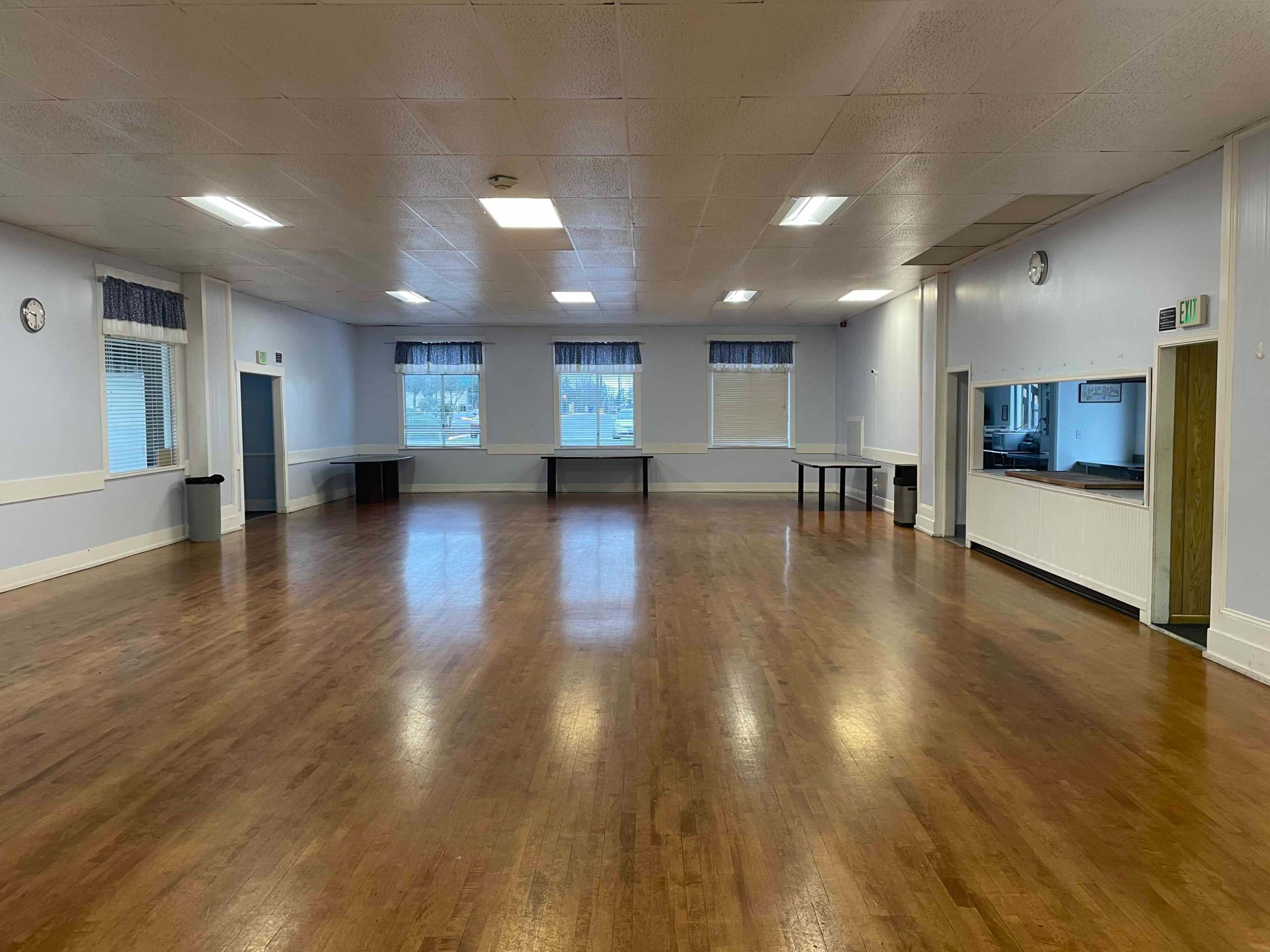 A spacious indoor hall with wood flooring, white walls, and large windows covered with blue curtains.