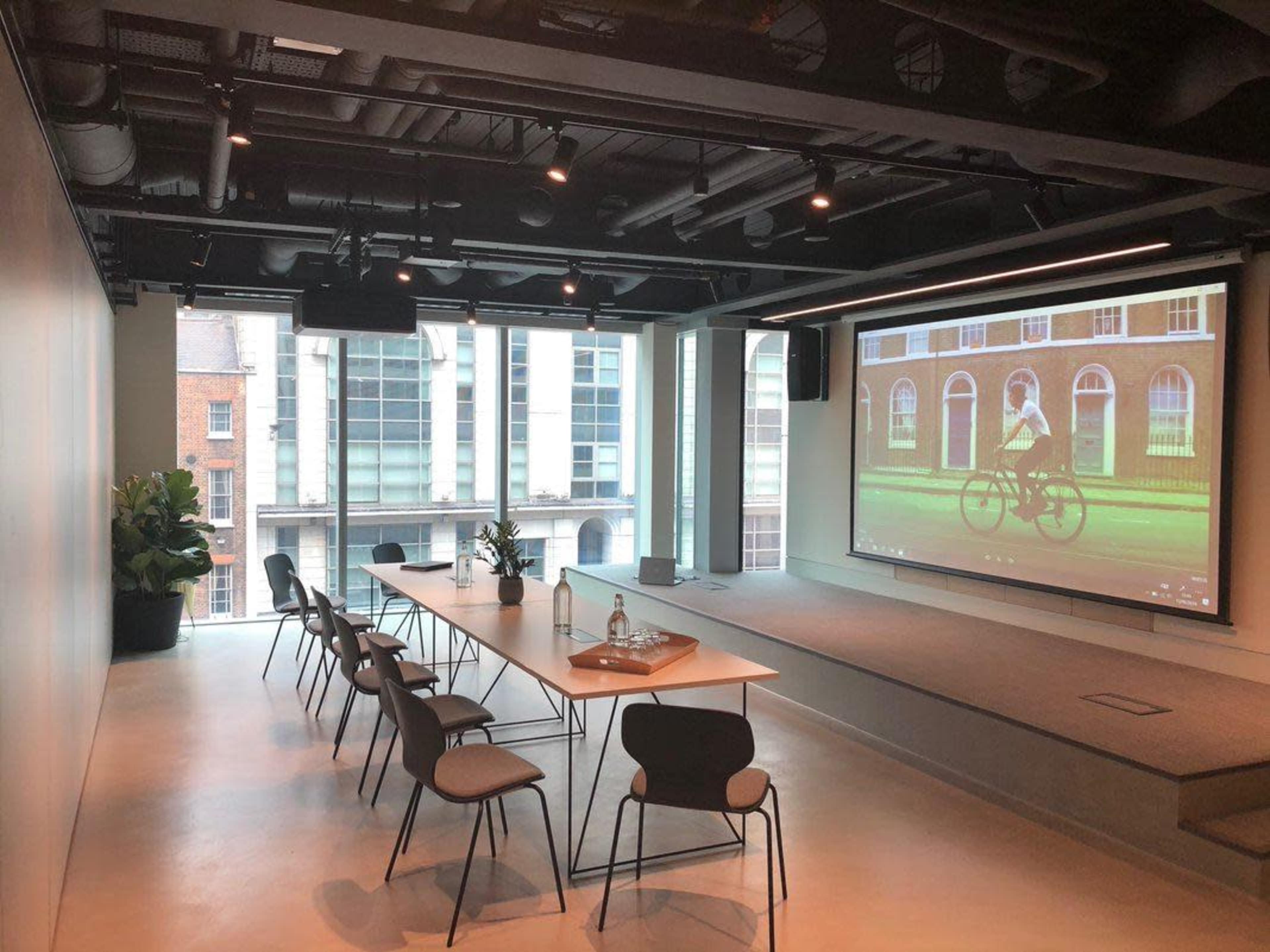 A modern conference room features a long table with chairs, large windows displaying a cityscape, and a projector screen showing a video.