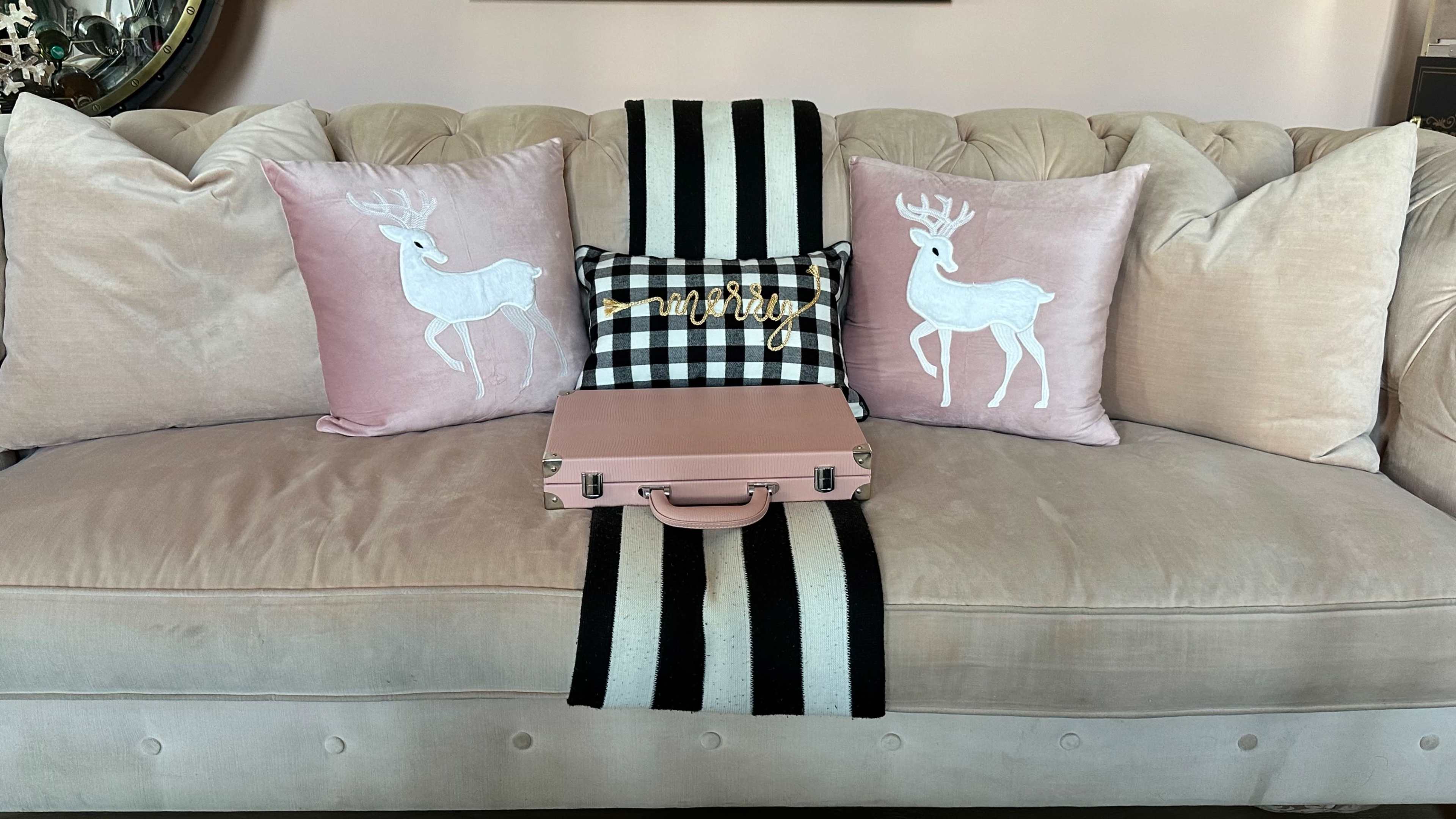 A pink velvet couch is adorned with decorative pillows featuring white deer and a black-and-white striped runner, with a pink suitcase placed on top.