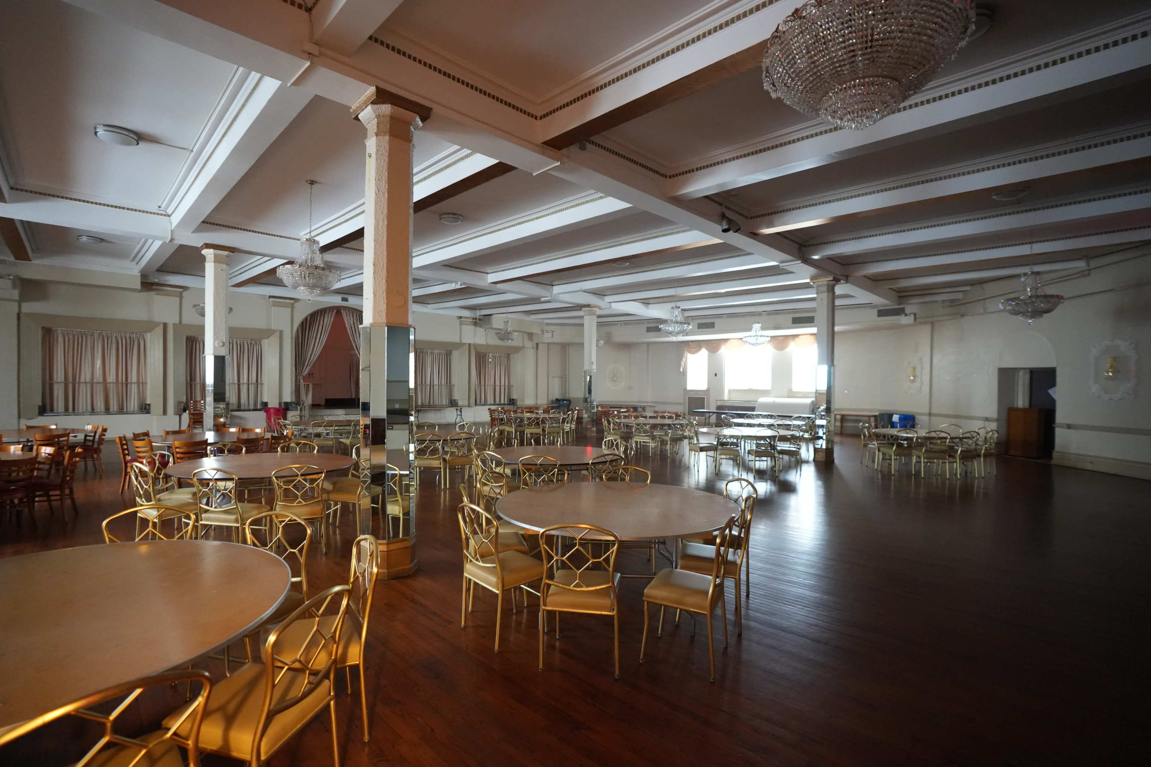 The image shows a large, empty hall featuring multiple round tables with gold chairs, polished wooden floors, and decorative ceiling beams.