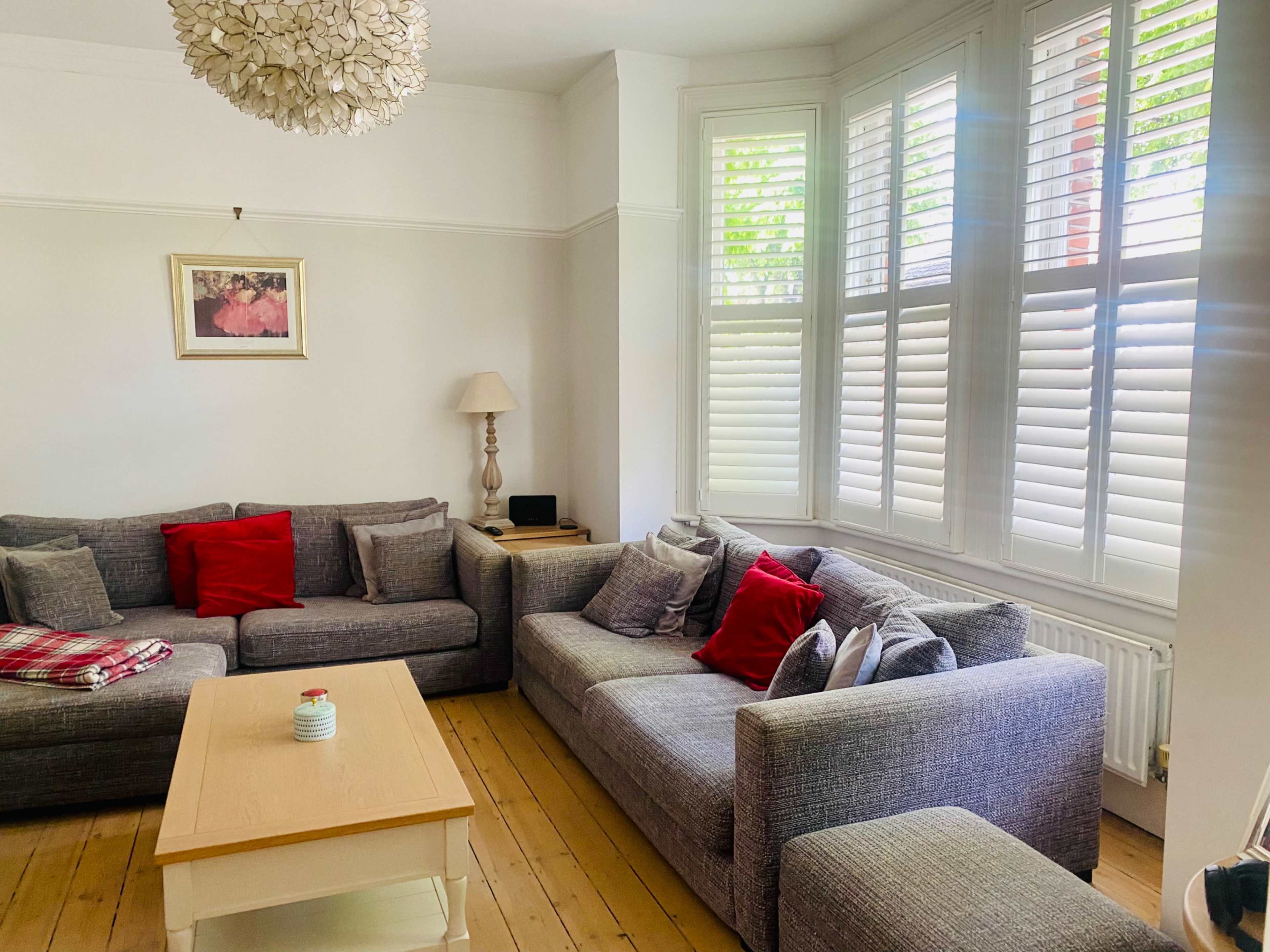 The image shows a cozy living room with a light-colored wooden floor, a grey sectional sofa adorned with red pillows, a wooden coffee table, and large windows dressed with white shutters.