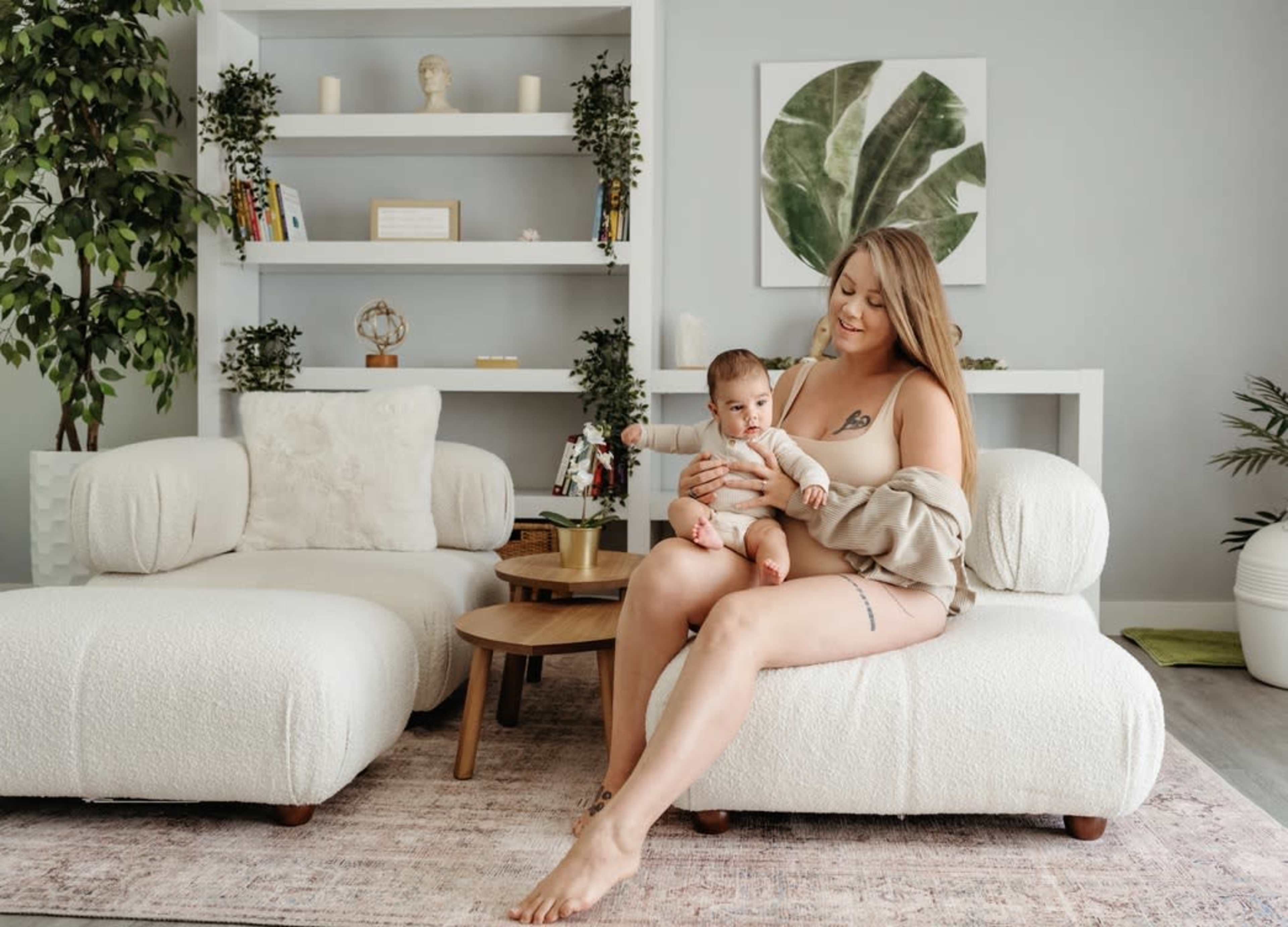 A woman holding a baby sits on a soft, upholstered chair in a modern, light-filled living room with plants and shelves in the background.