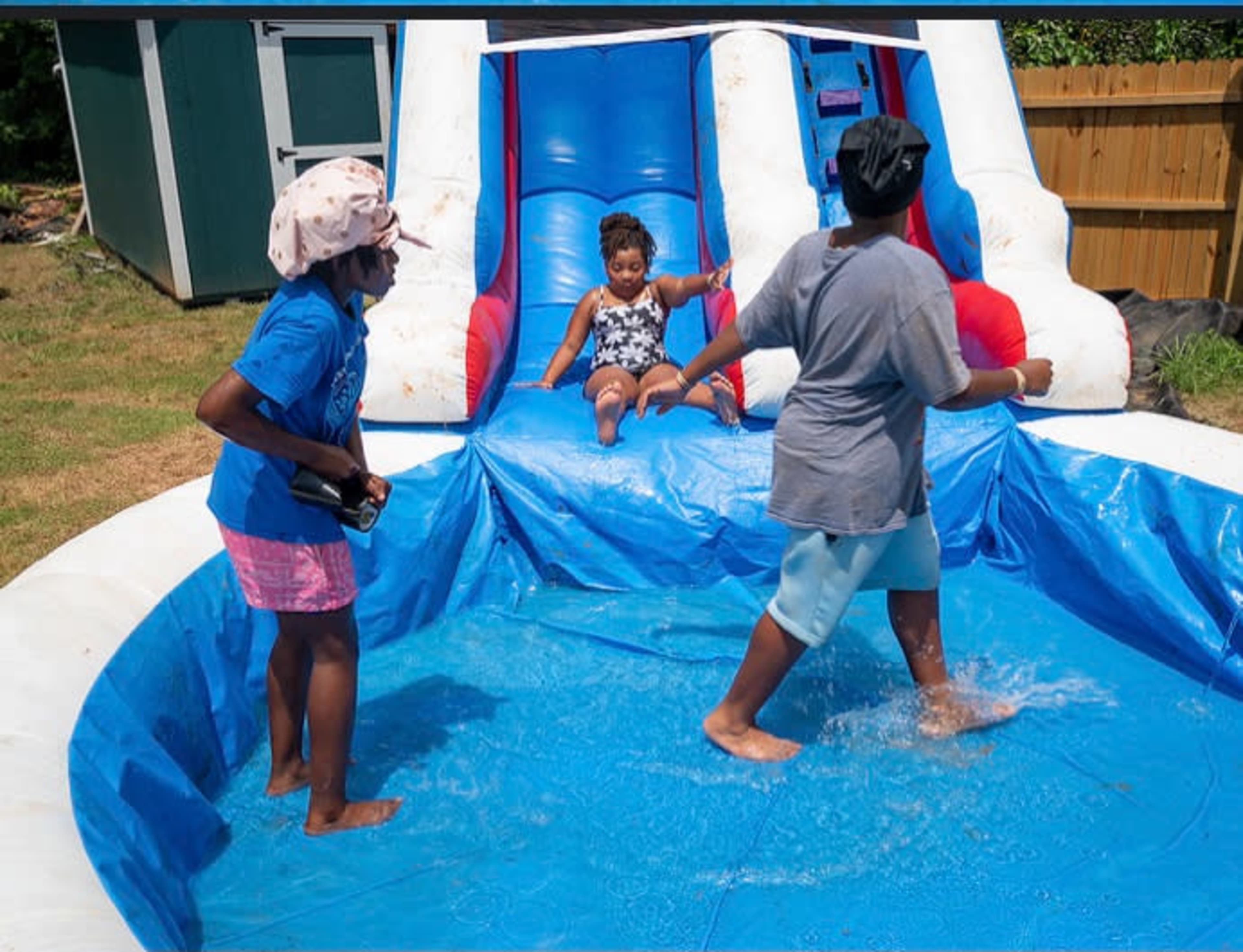Children are playing in a pool area with a water slide, while one child slides down and two others are standing in the water.