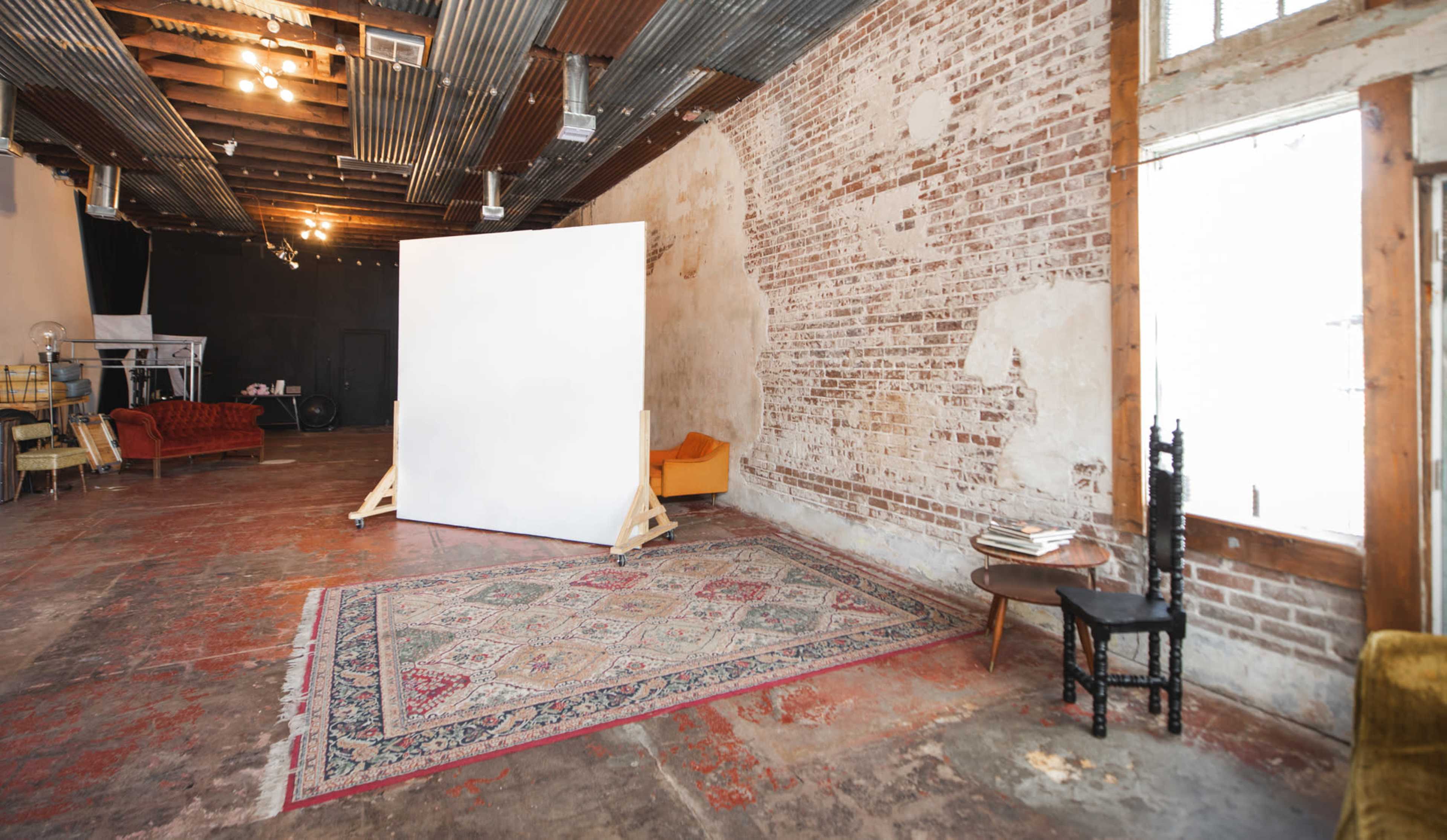 An industrial-style studio with exposed brick walls, a large white backdrop, and a colorful area rug on a concrete floor.