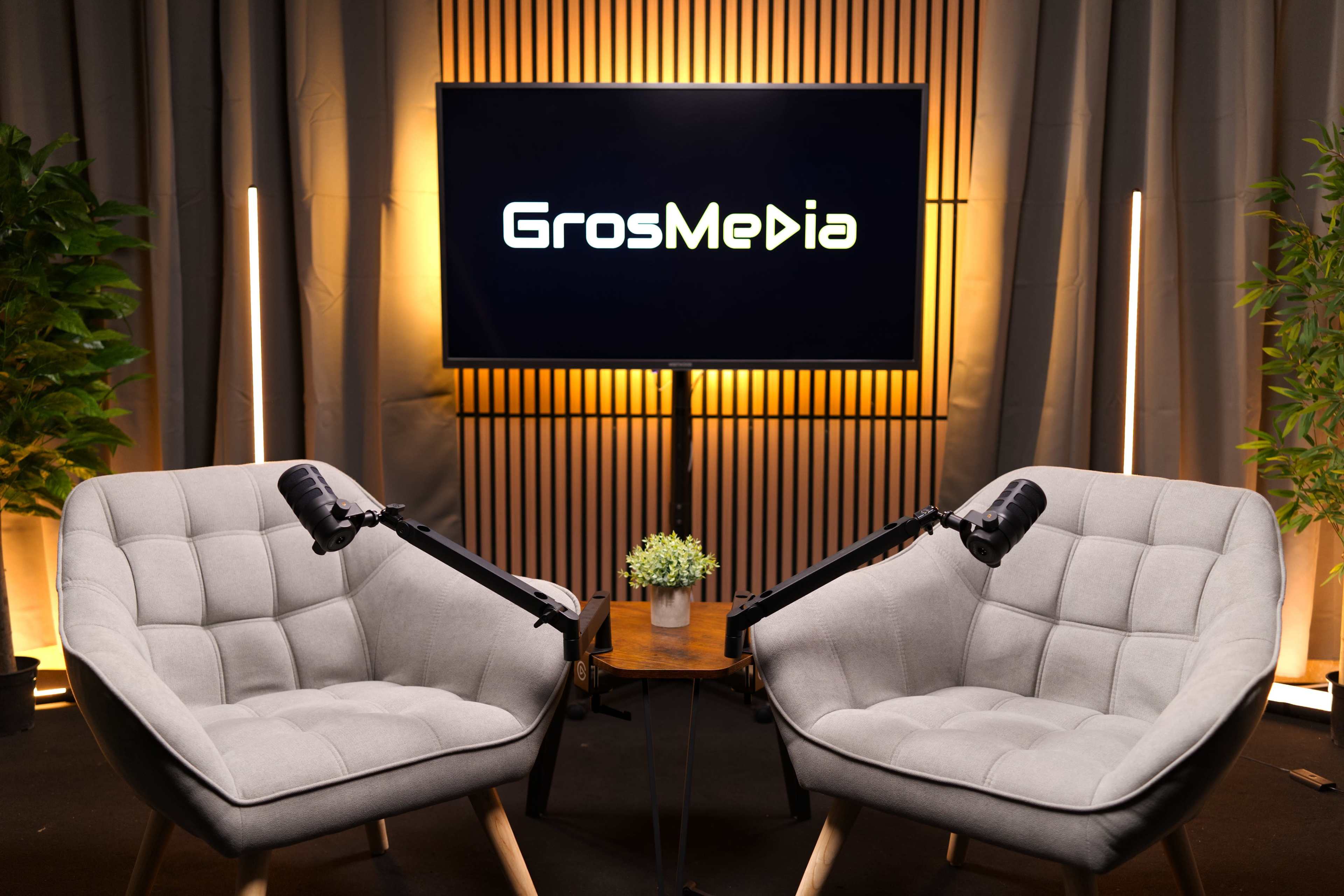 The image shows two gray armchairs with microphones on stands positioned beside a small table, set against a backdrop featuring a screen displaying the logo "GrosMedia."