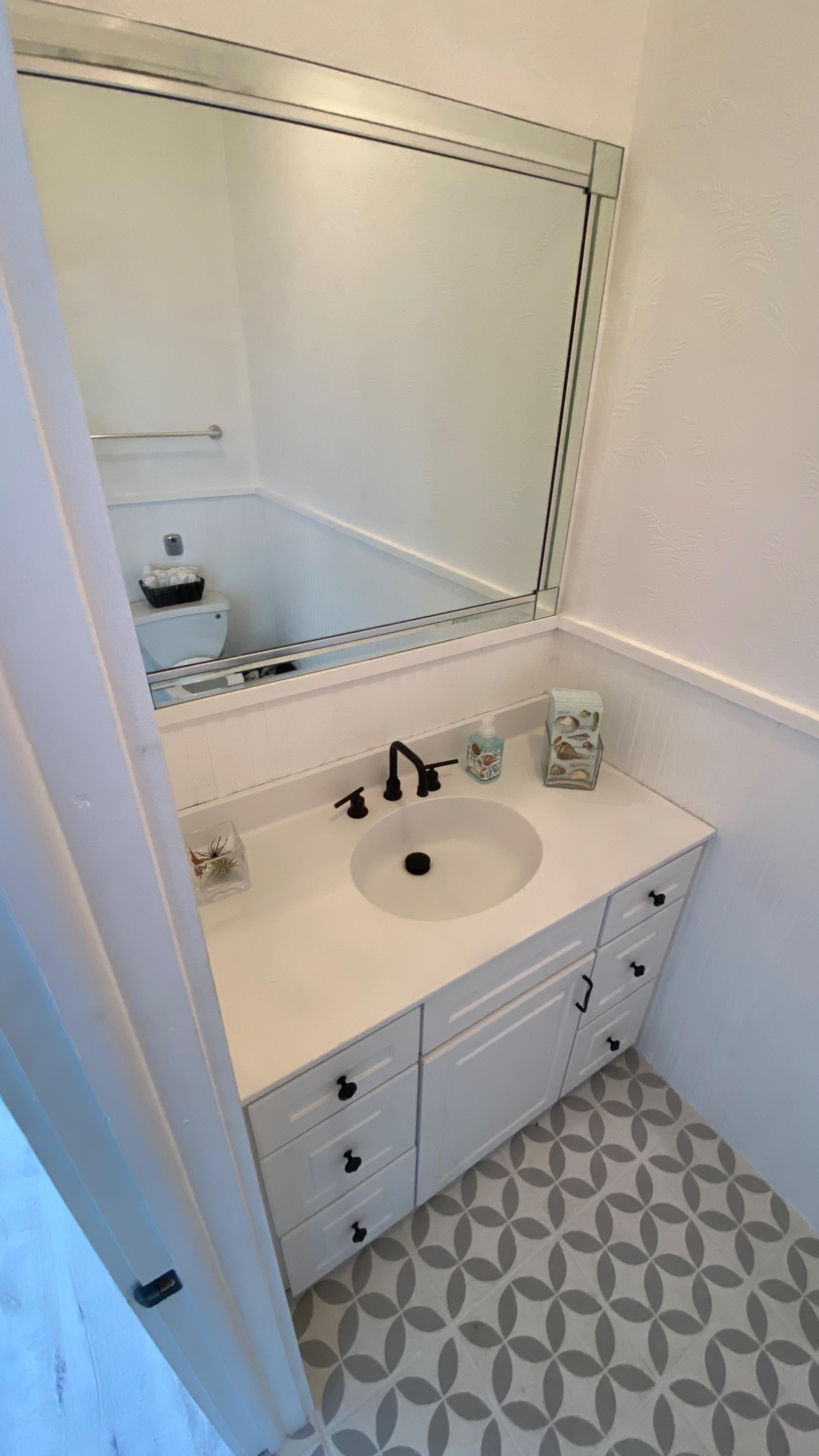 A small bathroom features a white vanity with a circular sink and a large mirror above it, surrounded by decorative items and a patterned tiled floor.
