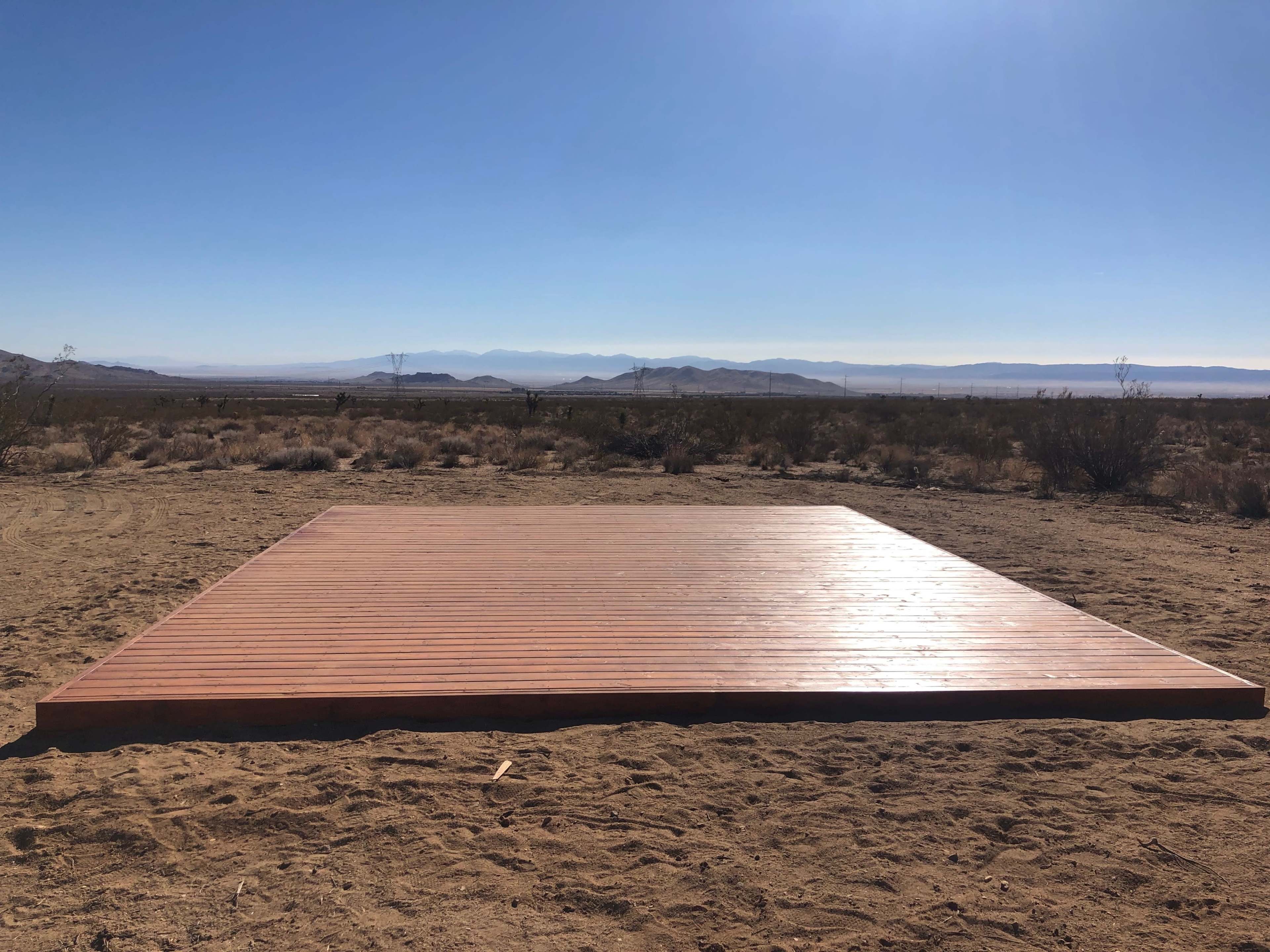 A large wooden platform is set on sandy ground in a desert landscape with mountains in the background under a clear blue sky.