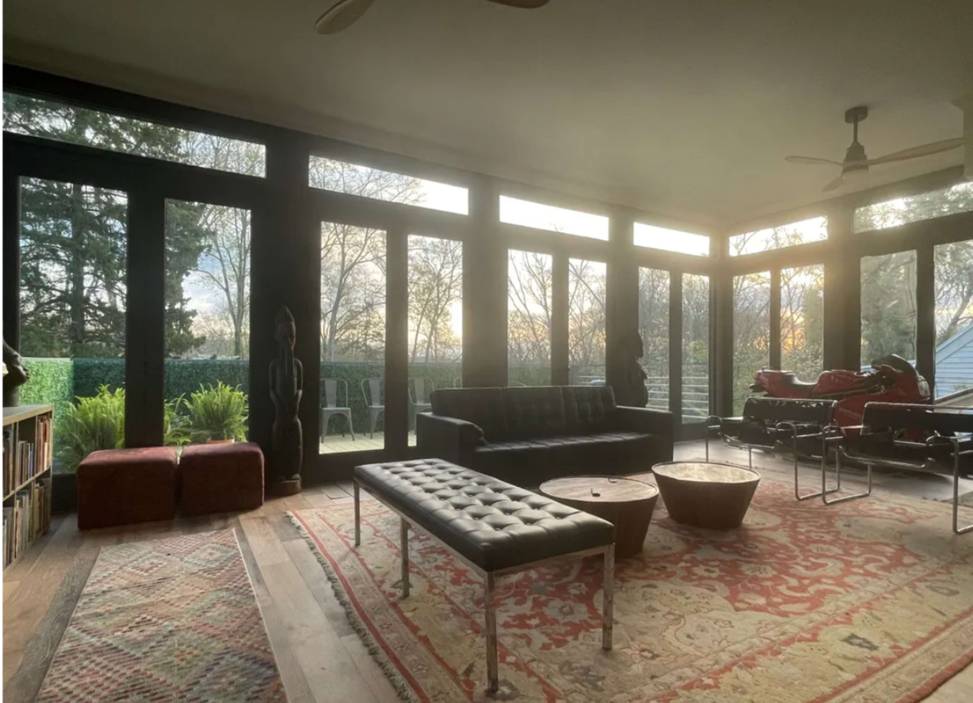 A sunlit living space with large windows, featuring a black sofa, a wooden coffee table, and a vintage motorcycle visible outside.