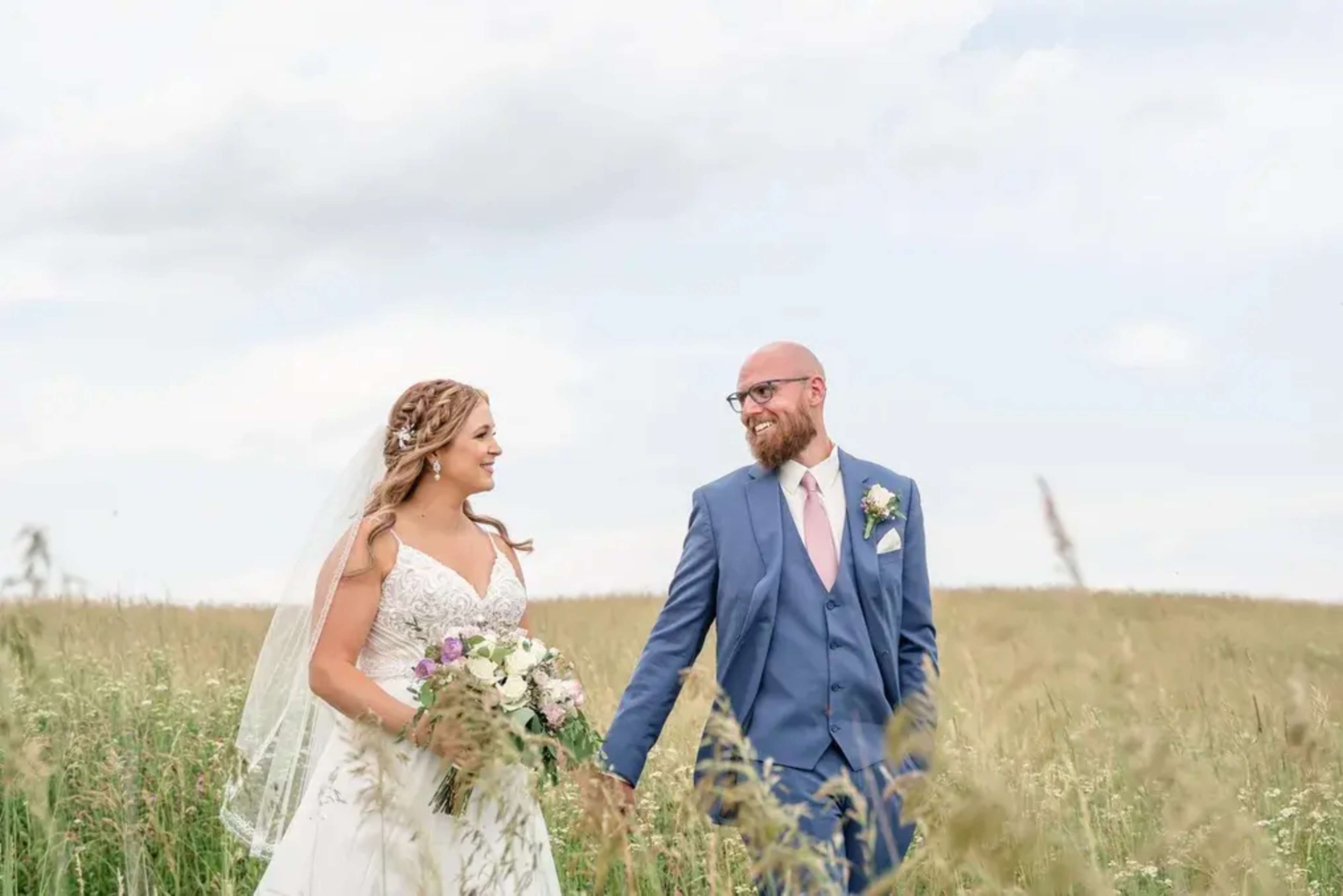 A bride and groom walk hand in hand through a field, smiling at each other.