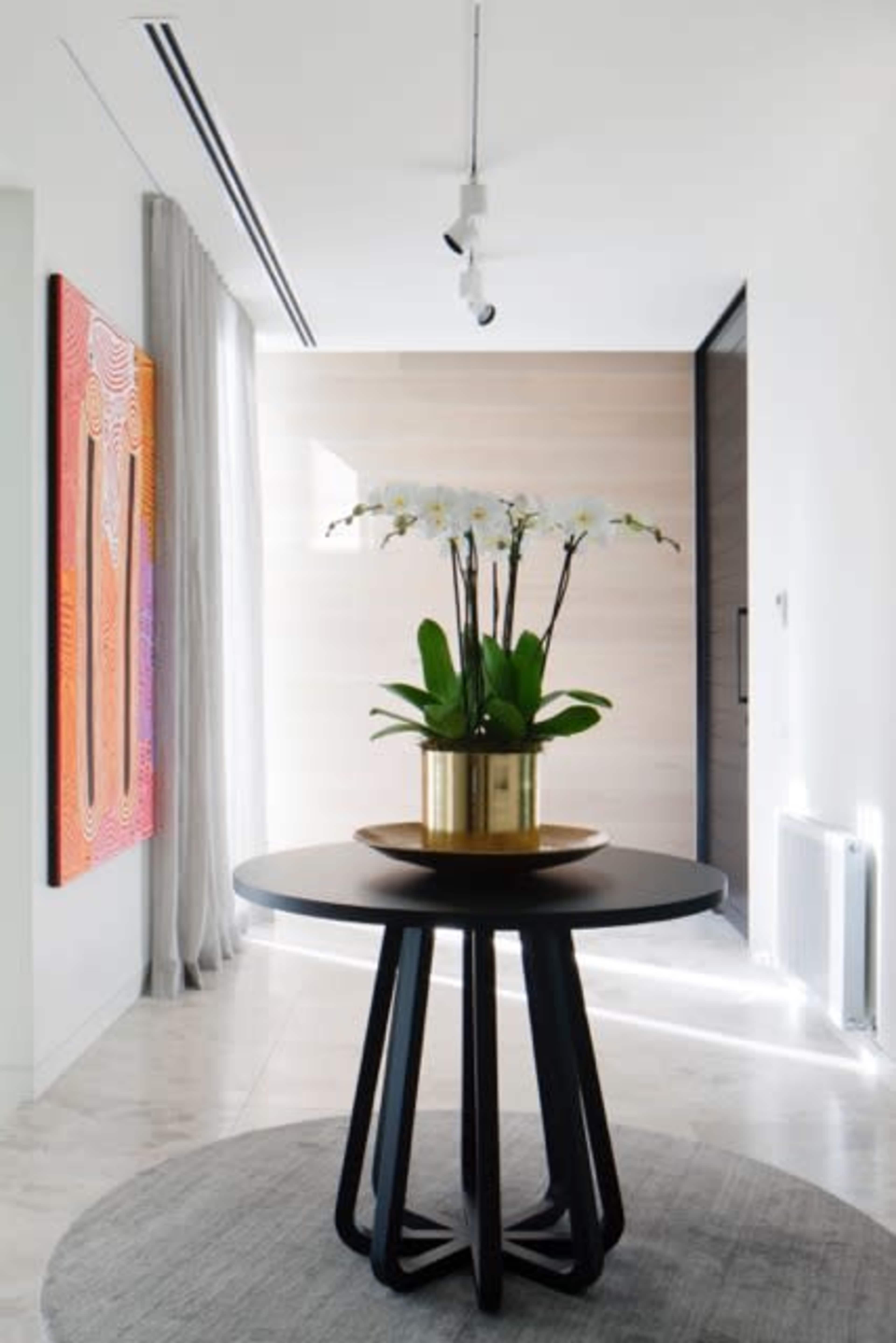 A black round table with a golden planter holding orchids stands in a well-lit hallway next to an abstract painting and sheer curtains.
