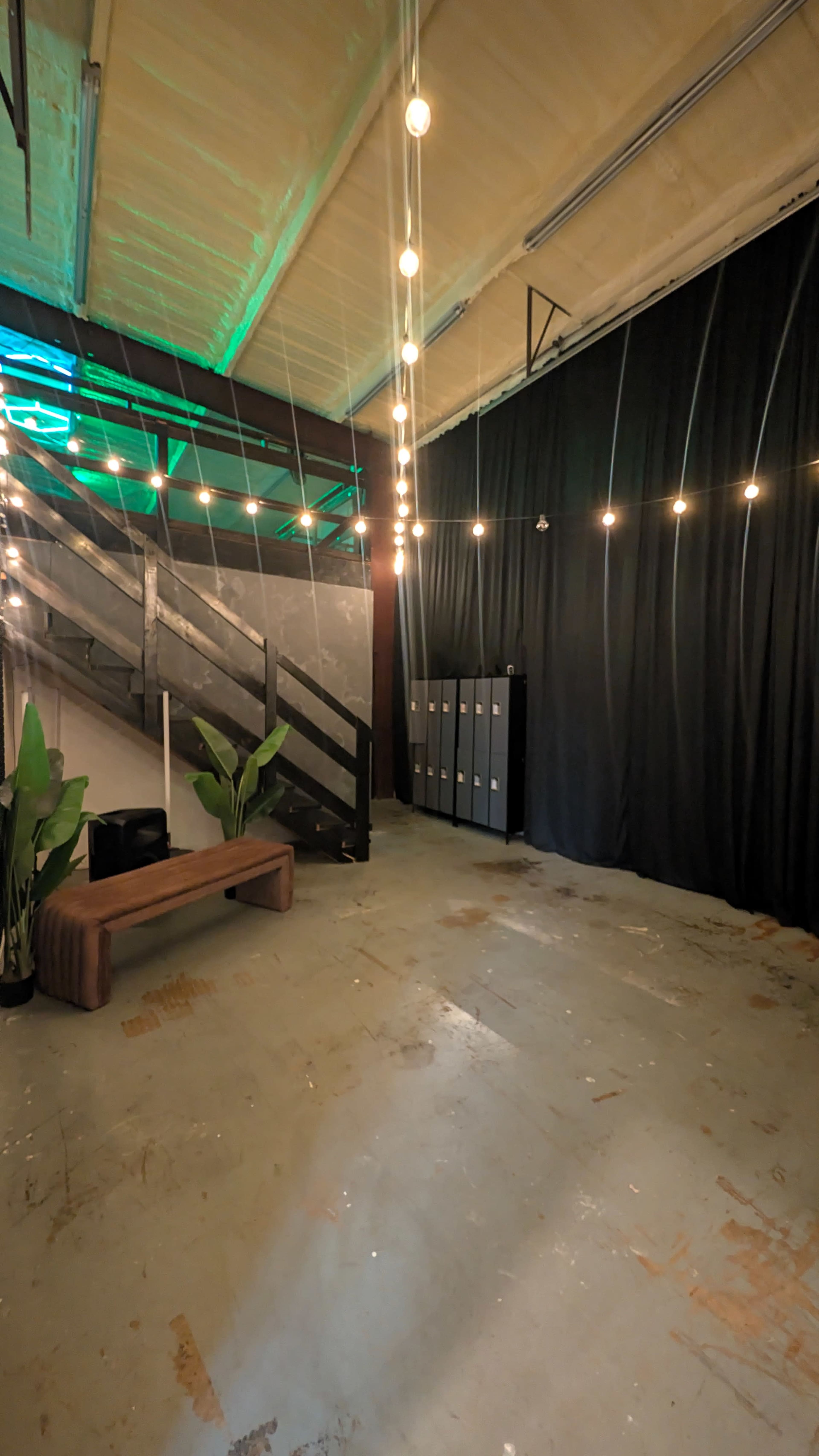 The image shows a spacious room with a wooden bench, decorative plants, lockers against a black curtain, and string lights overhead.