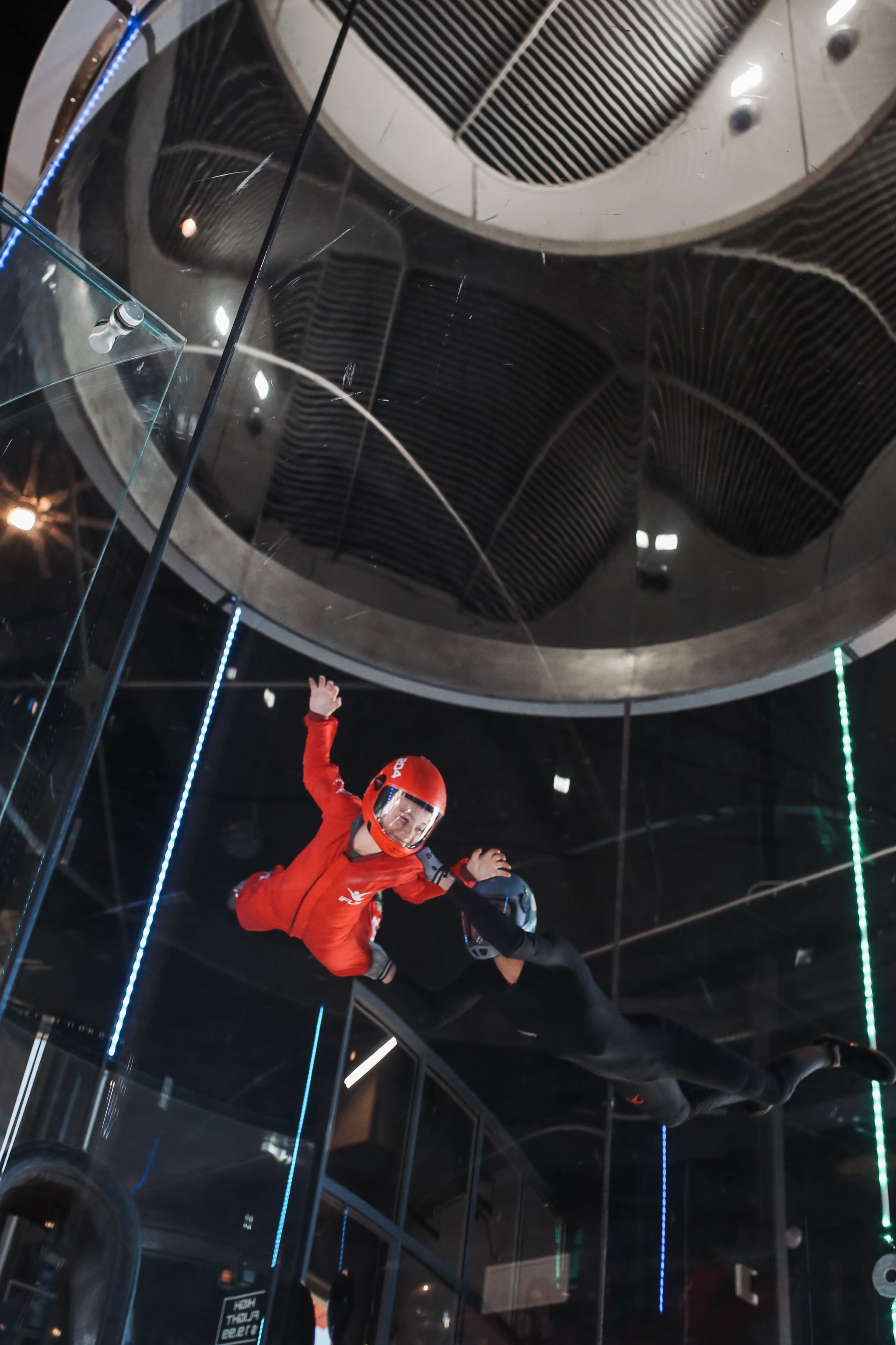 Two individuals in jumpsuits are performing indoor skydiving inside a vertical wind tunnel.