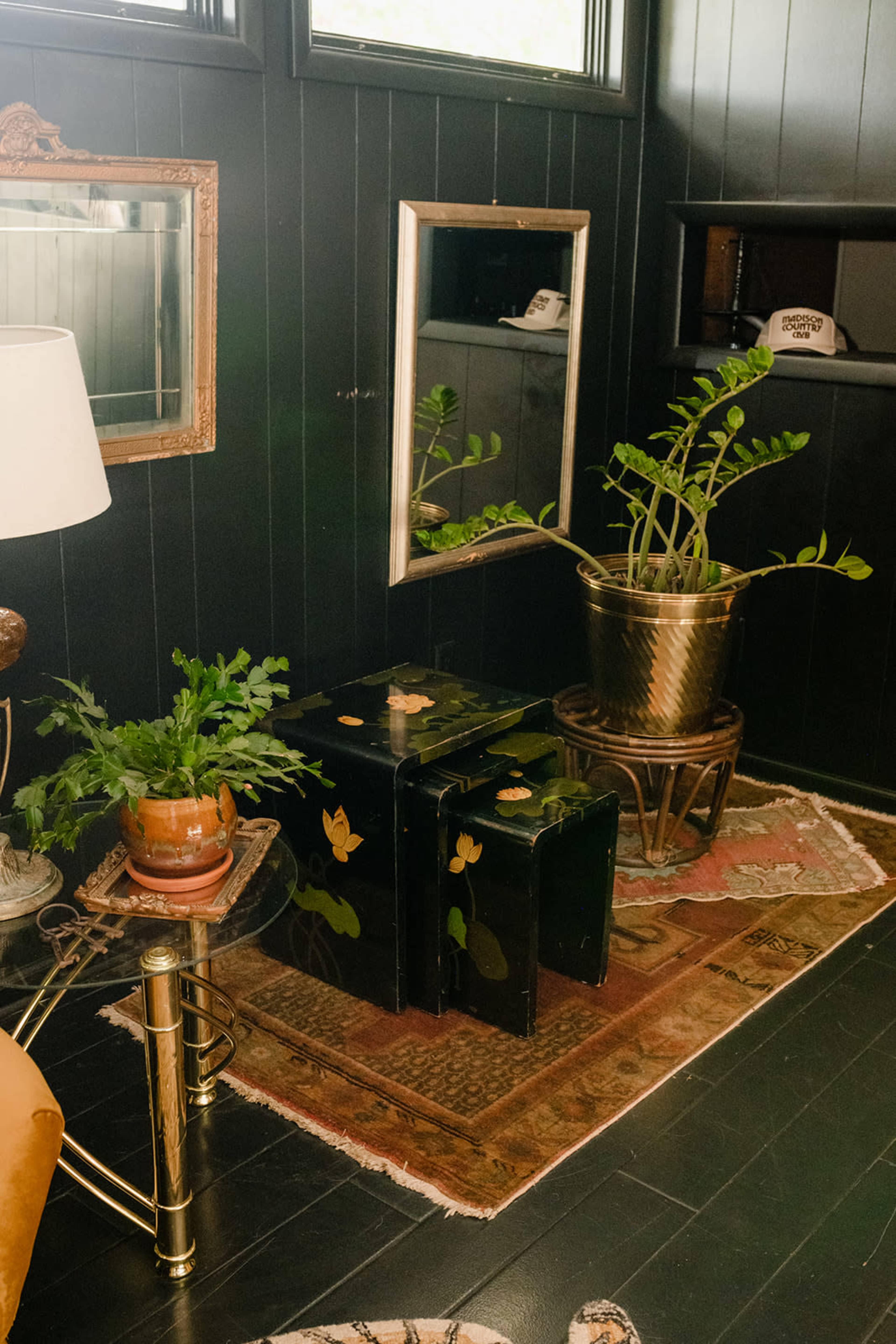A corner of a room with dark wooden walls, a small table with a lamp, a black box with decorative motifs, a potted plant, and a gold-colored planter on a stand.
