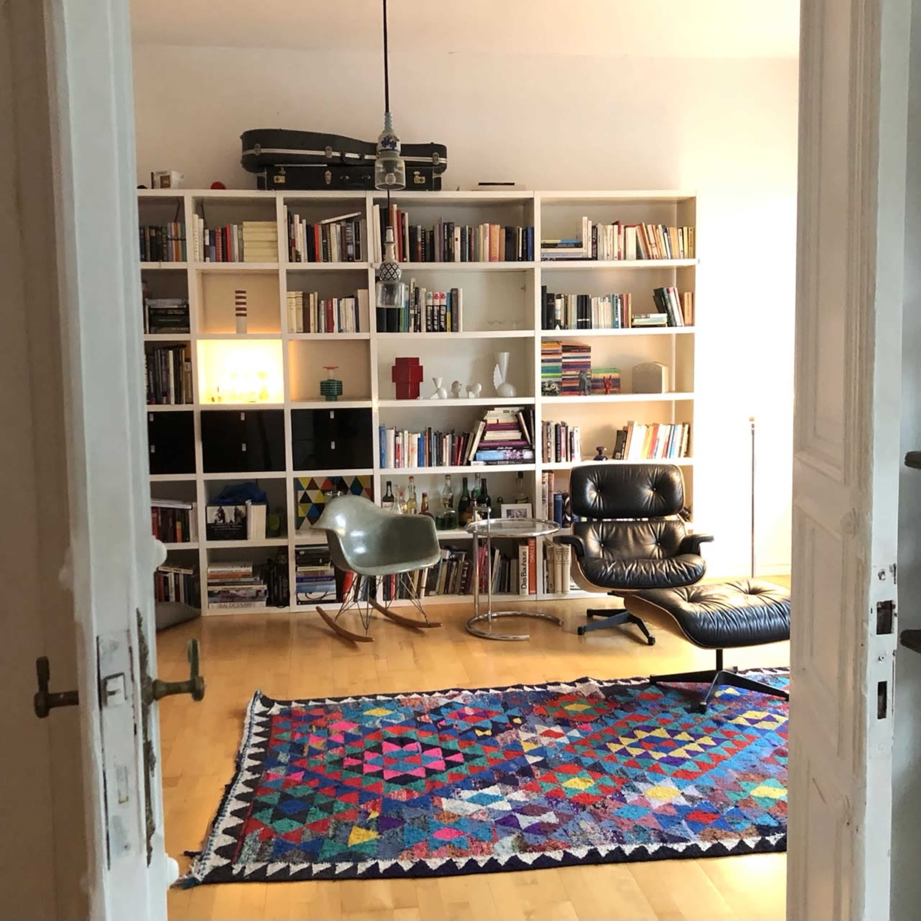 A bright room features a bookshelf filled with books, a lounge chair, and a colorful rug on a wooden floor.