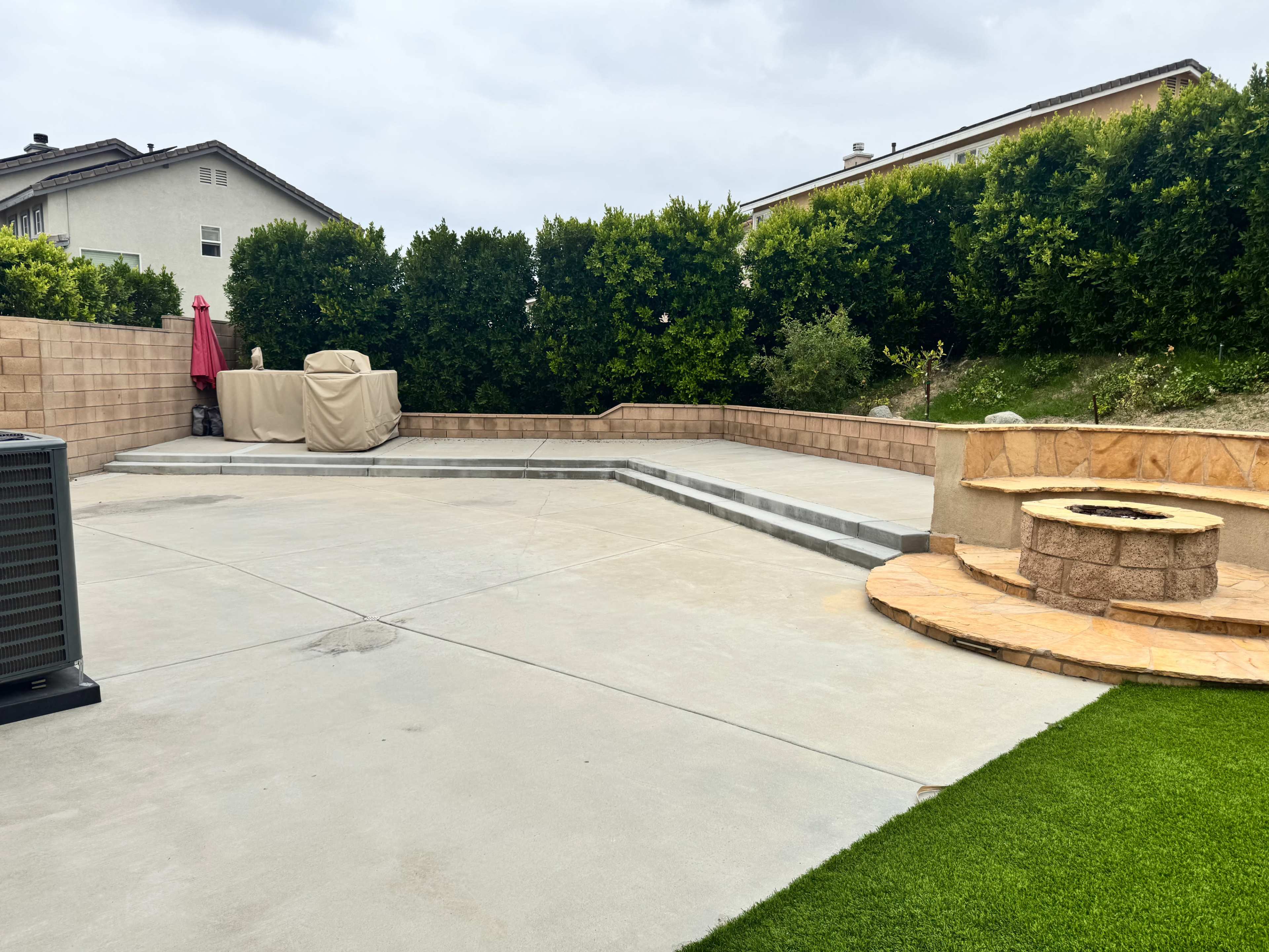 The image shows a spacious outdoor patio area with a stone fire pit, a covered grill, and a green lawn in a residential setting surrounded by hedges.