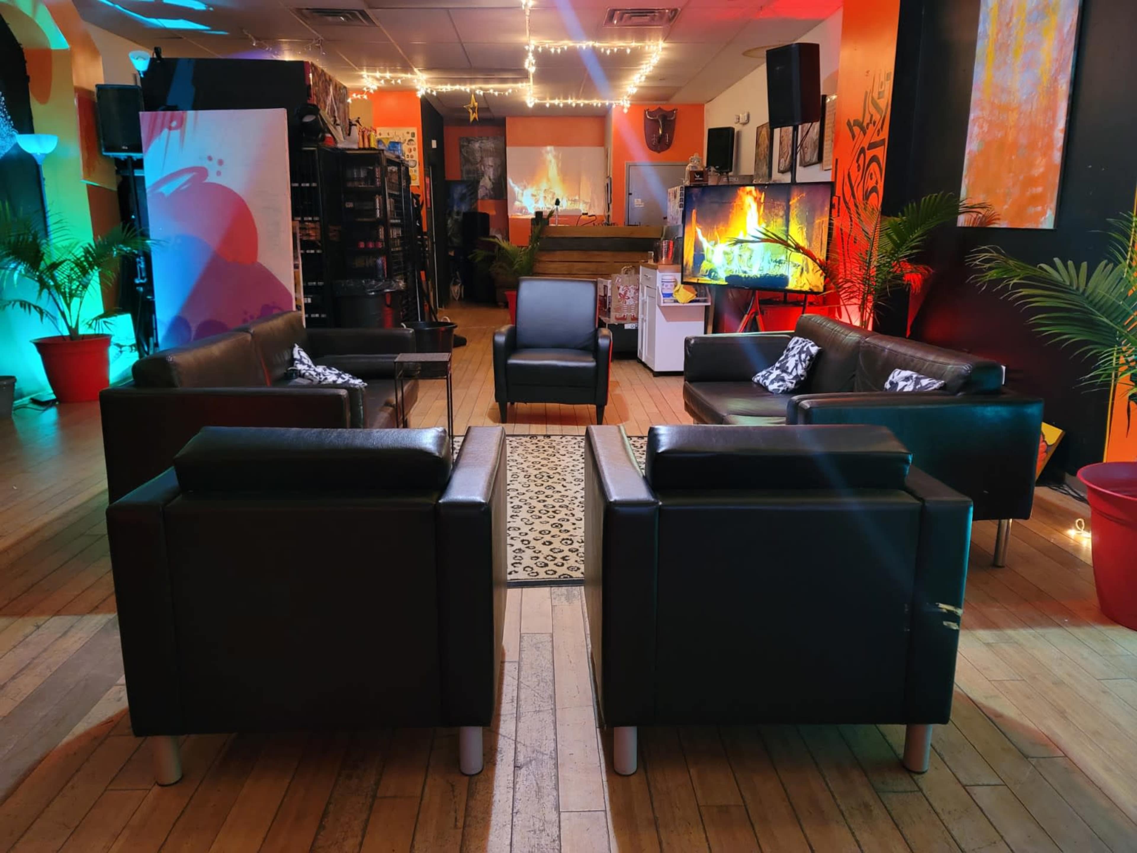 The image shows a cozy lounge area with black leather sofas arranged around a central rug, illuminated by colorful lights against a backdrop of art-covered walls.