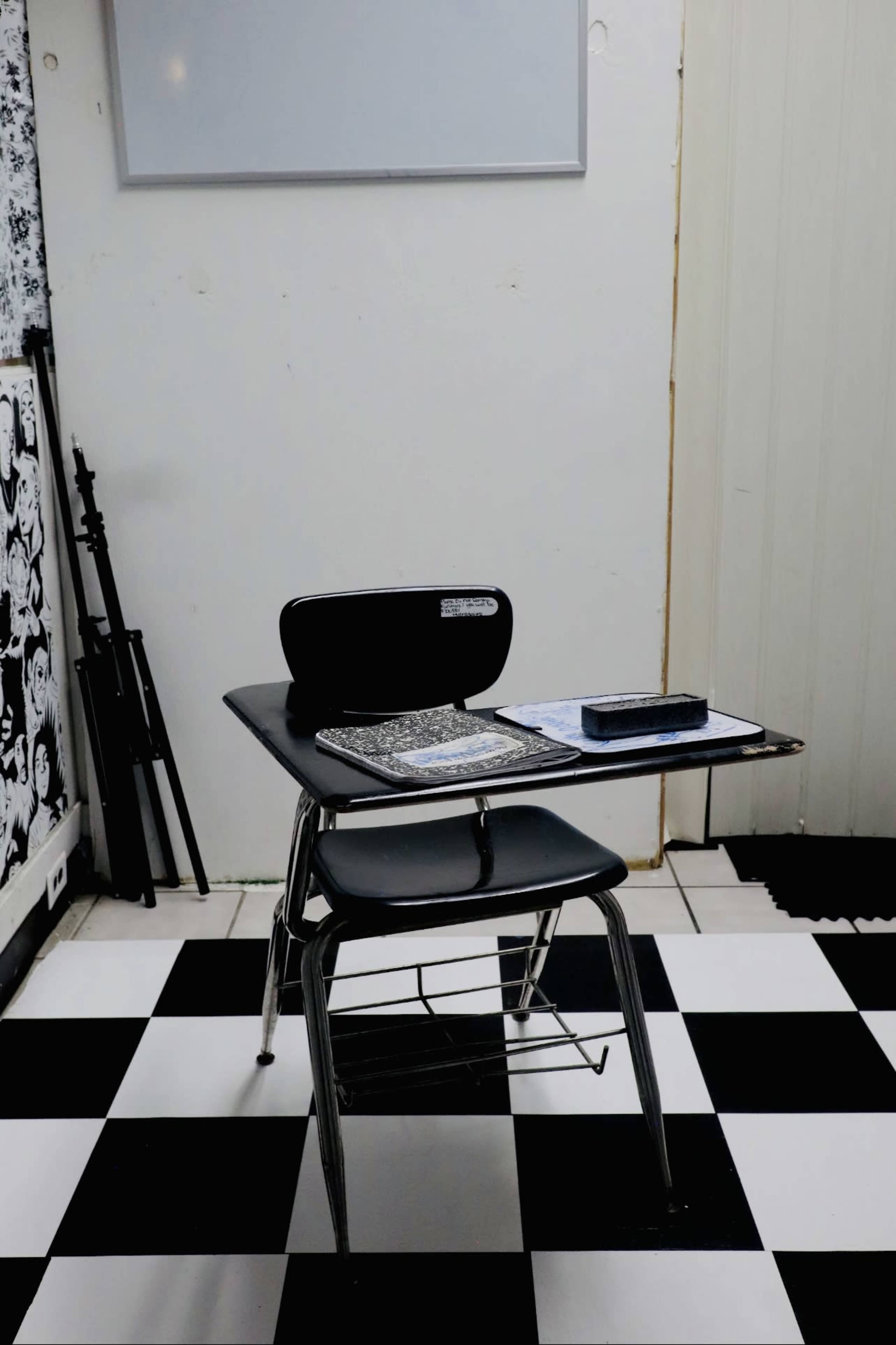 A single black and metal school desk with a notebook and a pencil rests on a black and white checkered floor, next to a blank whiteboard.