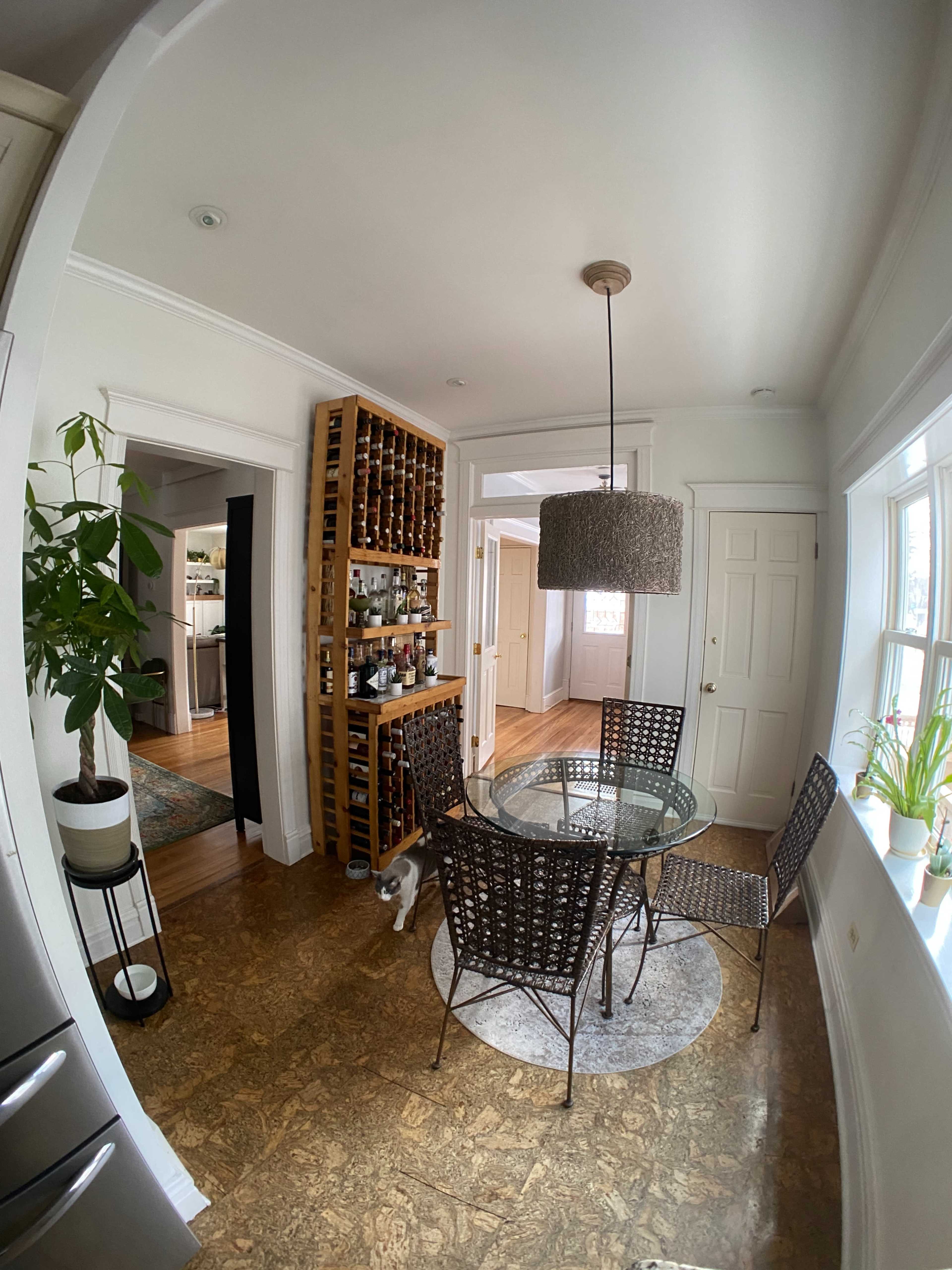 A small dining area features a round glass table surrounded by four wicker chairs, a wine rack filled with bottles, and a plant in the corner.