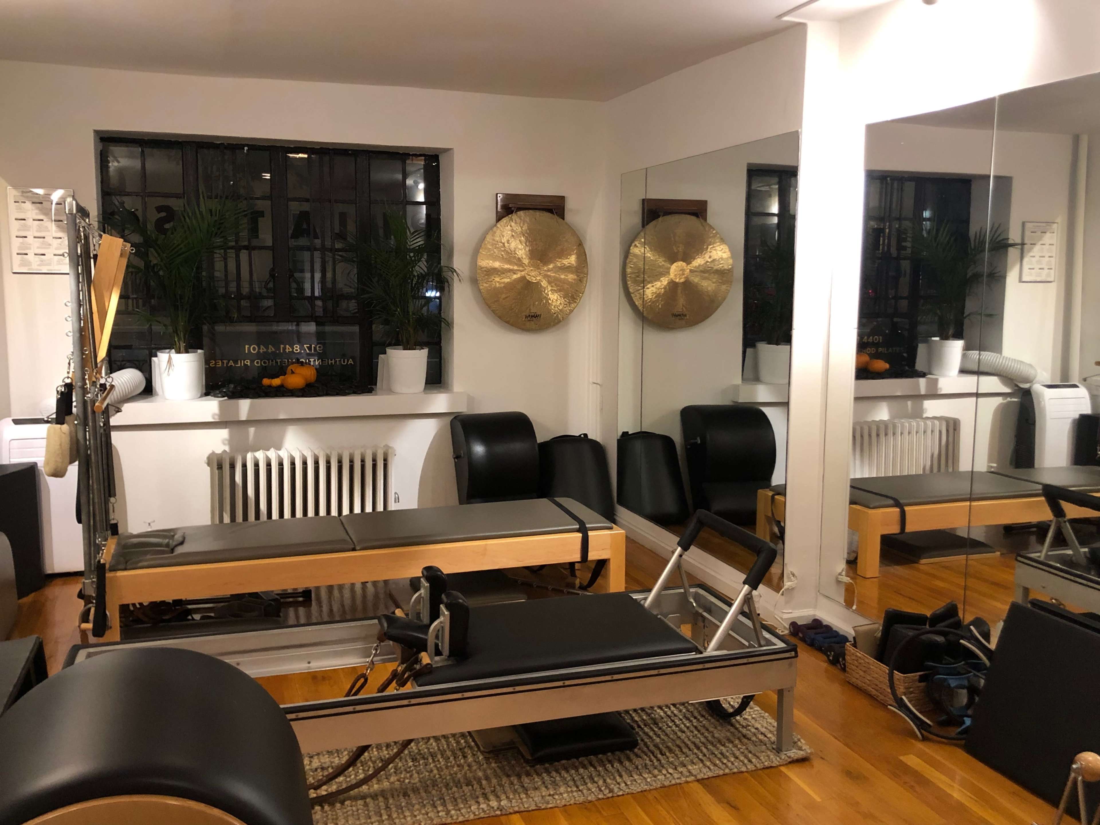 A Pilates studio featuring a reformer, large mirrors, and potted plants, with a decorative wall including a gong and a radiator.