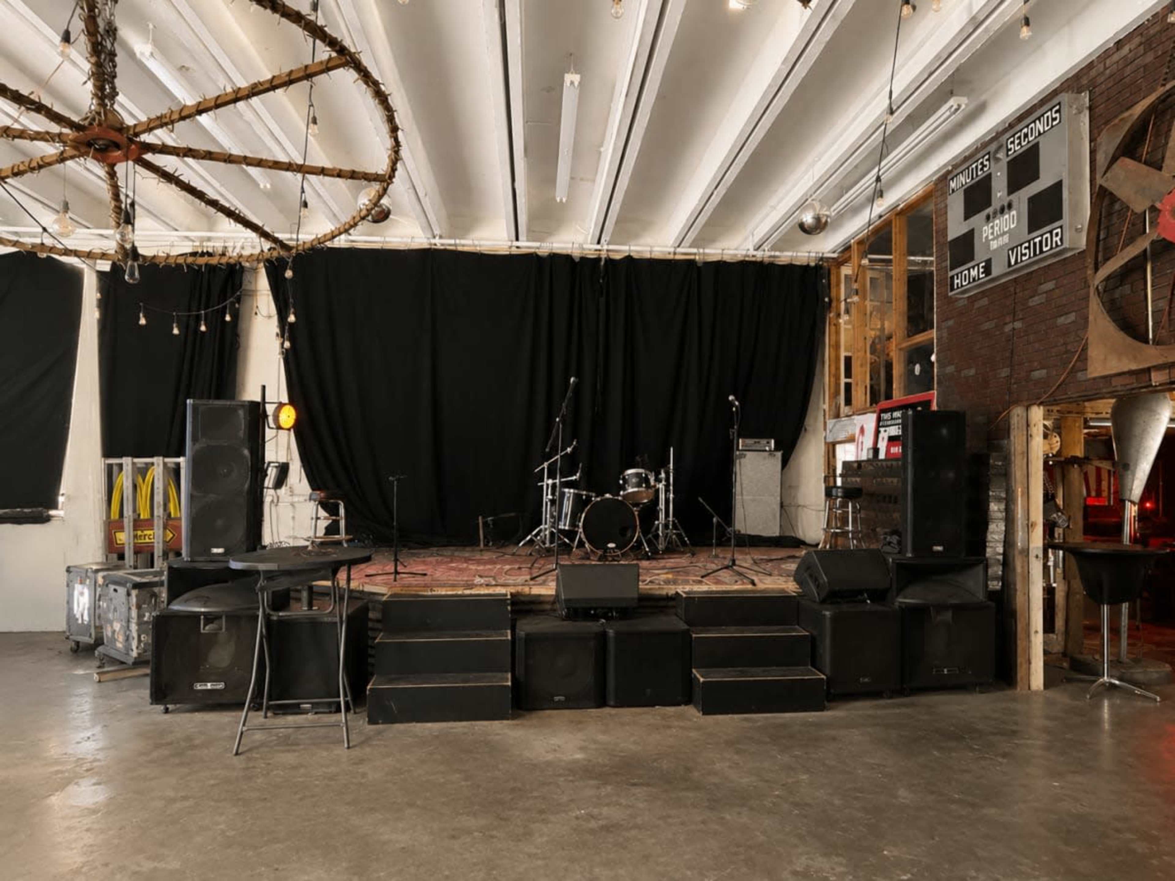 The image shows a stage with musical equipment, including drum sets and speakers, in a venue set up for performances, with a black curtain backdrop.