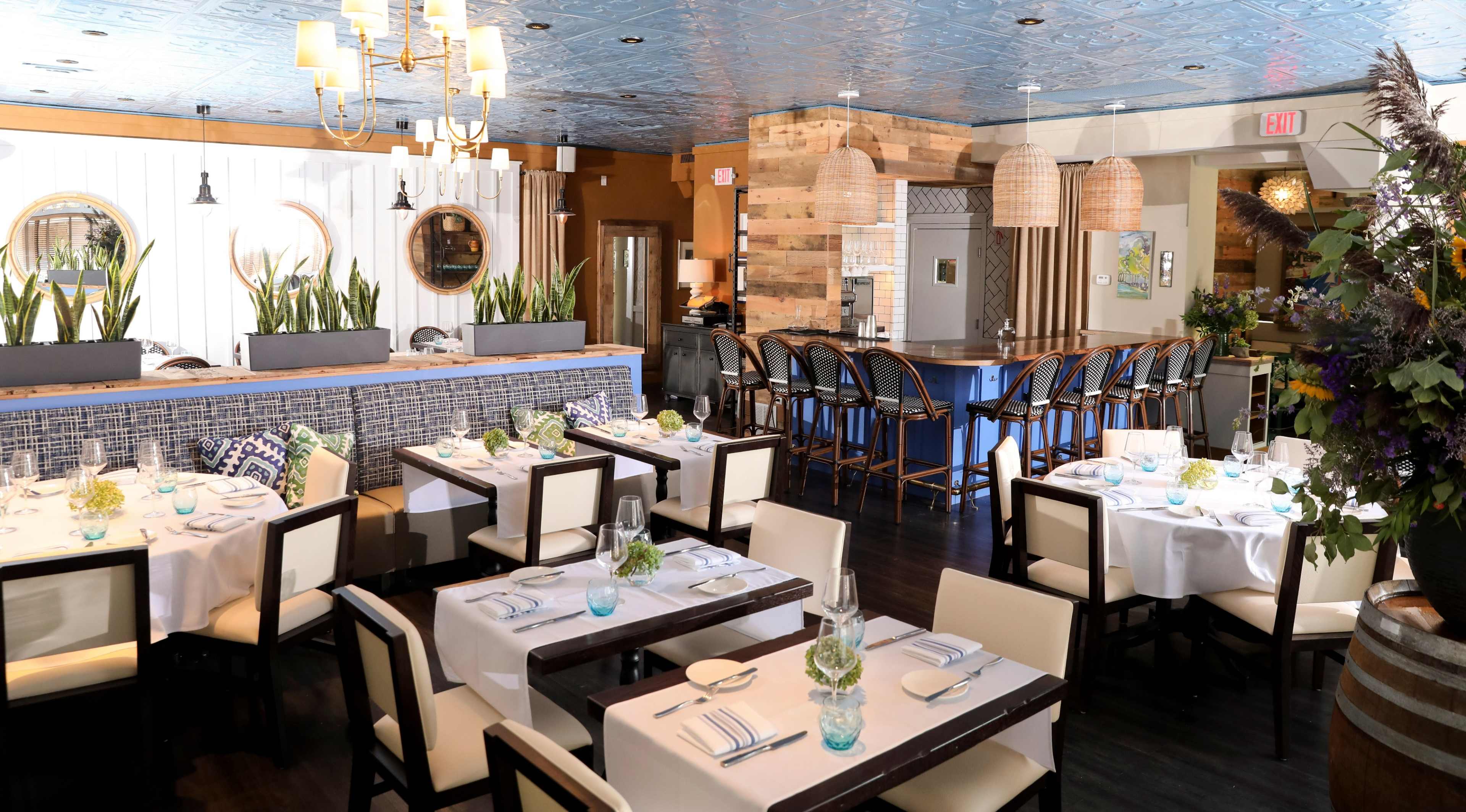A modern restaurant interior with wooden tables, white tablecloths, and a blue ceiling, featuring a bar area and potted plants.