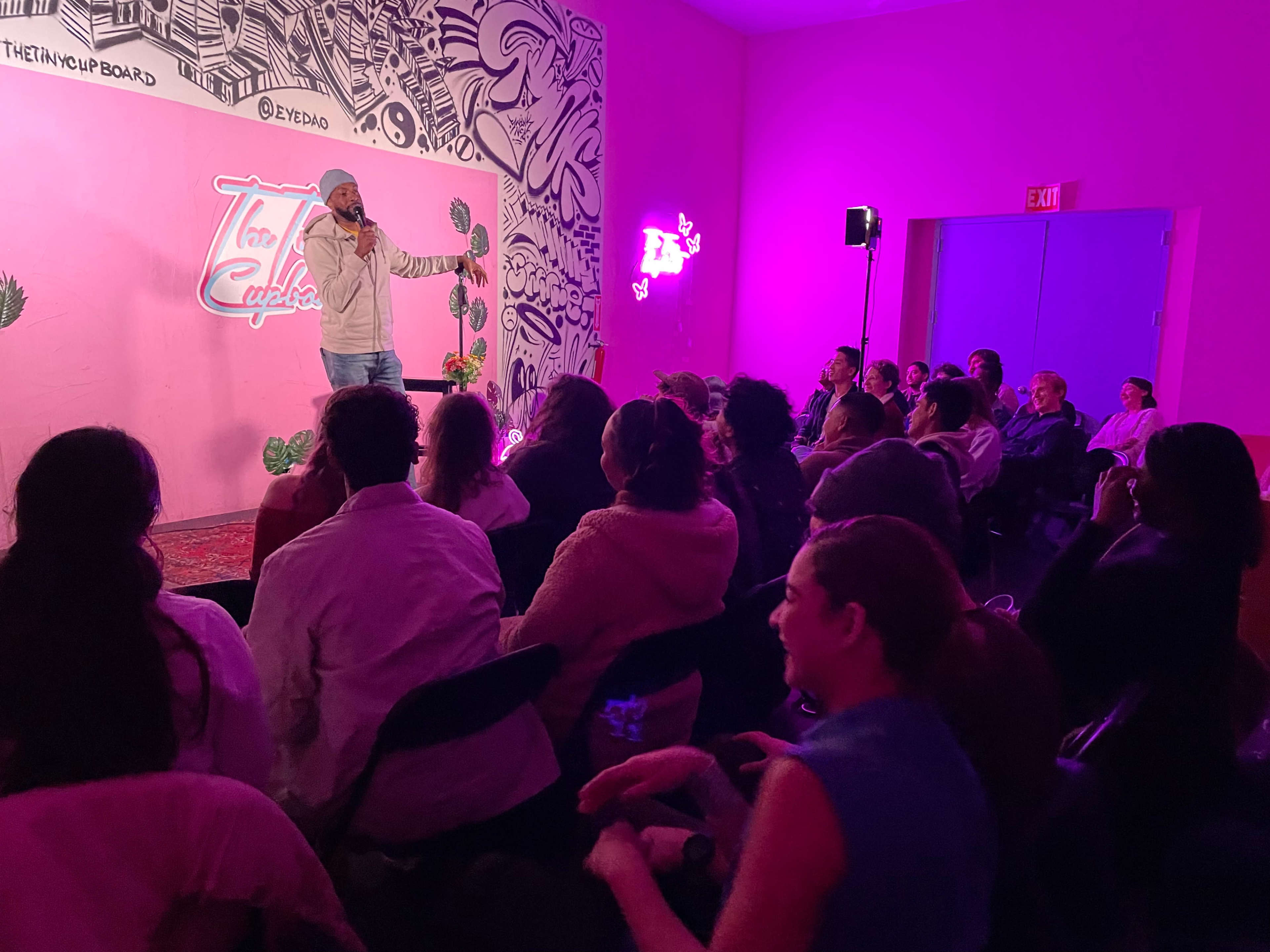A performer stands on stage speaking to an audience seated in a brightly colored room adorned with decorative elements.