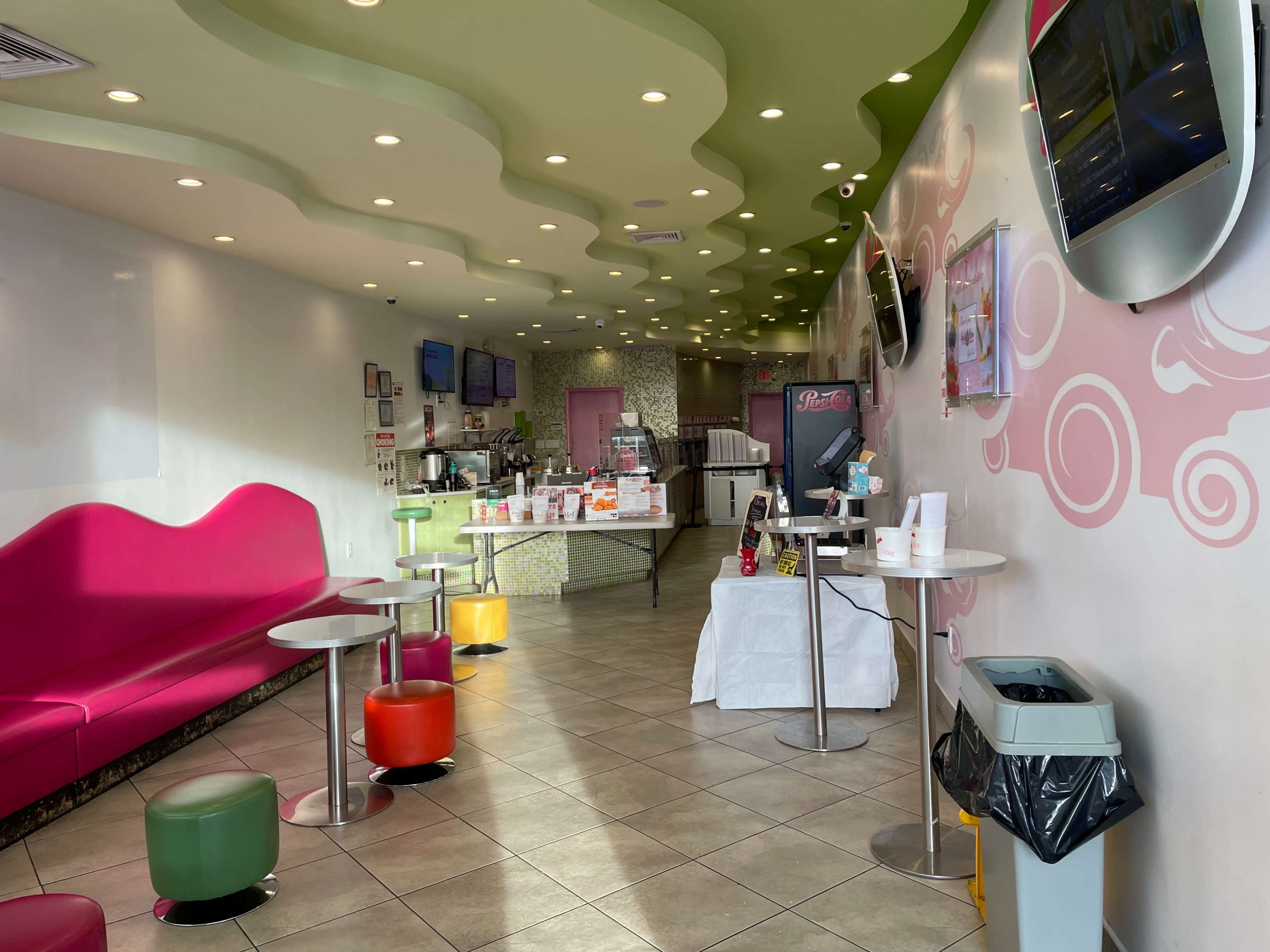 A brightly colored dessert shop interior with a pink seating area, circular tables, and a serving counter visible at the back.