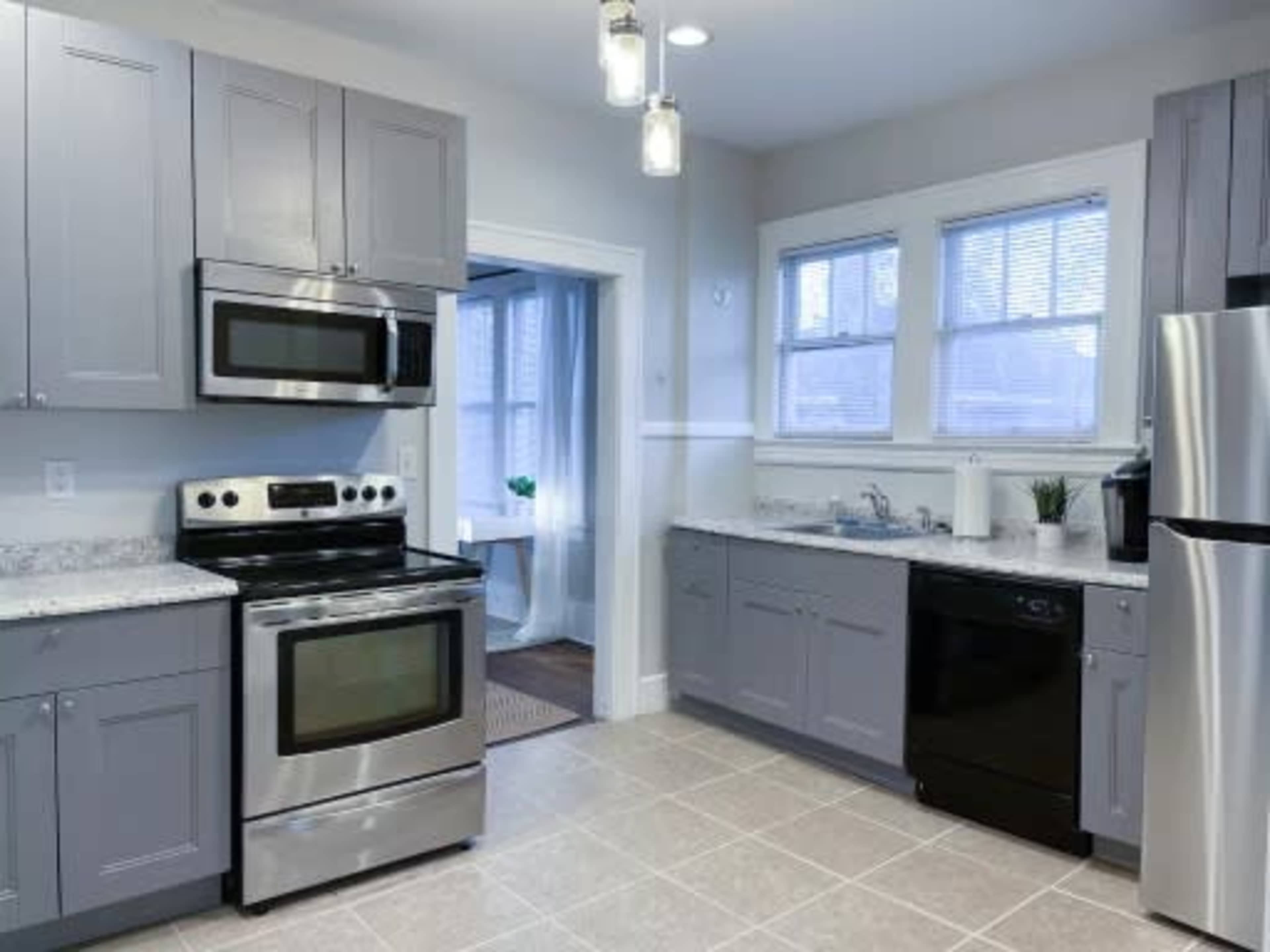 The kitchen features gray cabinets, stainless steel appliances, and tiled flooring with a view into a well-lit adjacent room.