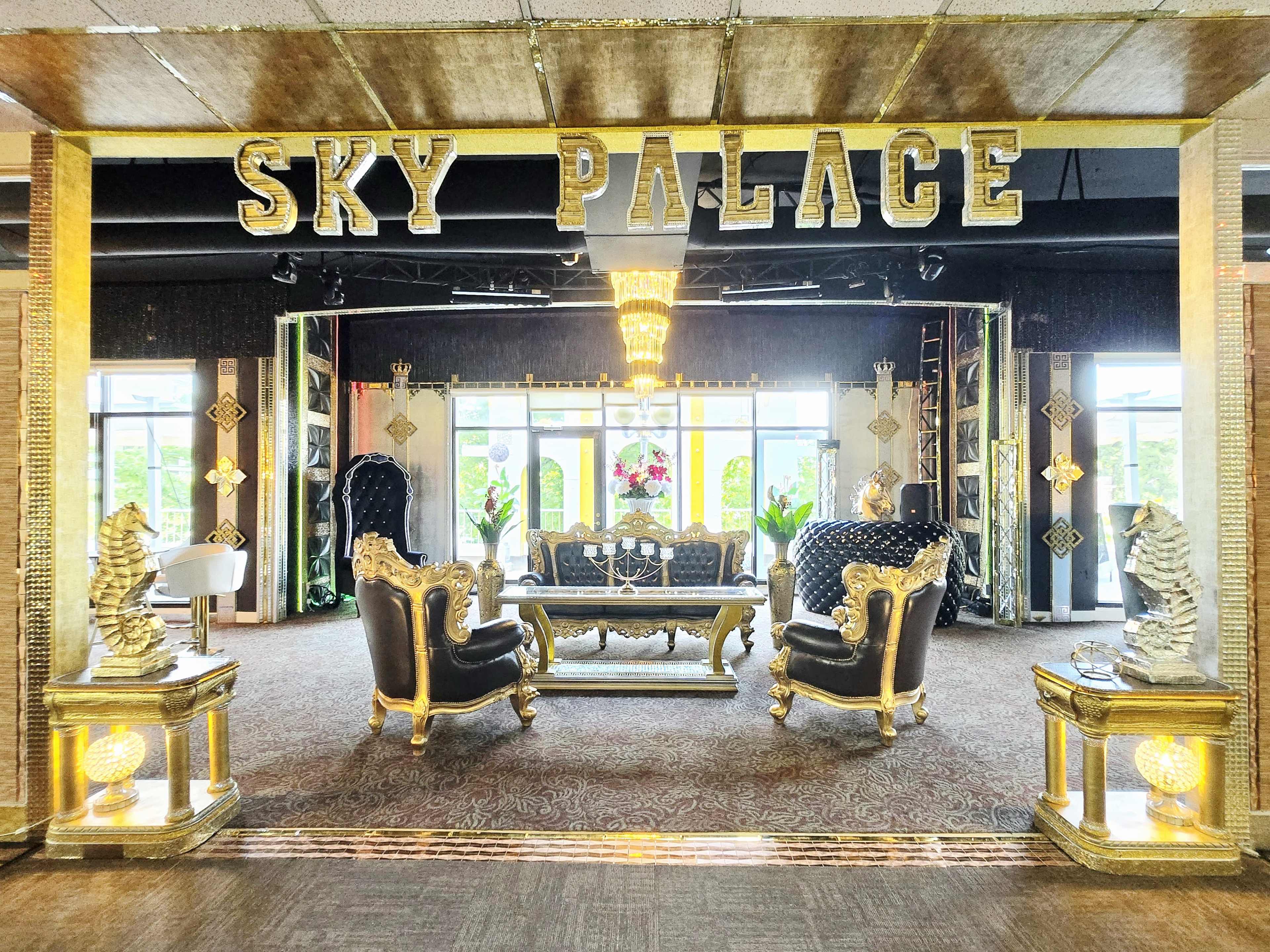 The image shows an elaborately decorated lounge area with ornate black and gold furniture, featuring large armchairs, a central coffee table, and a grand chandelier above.