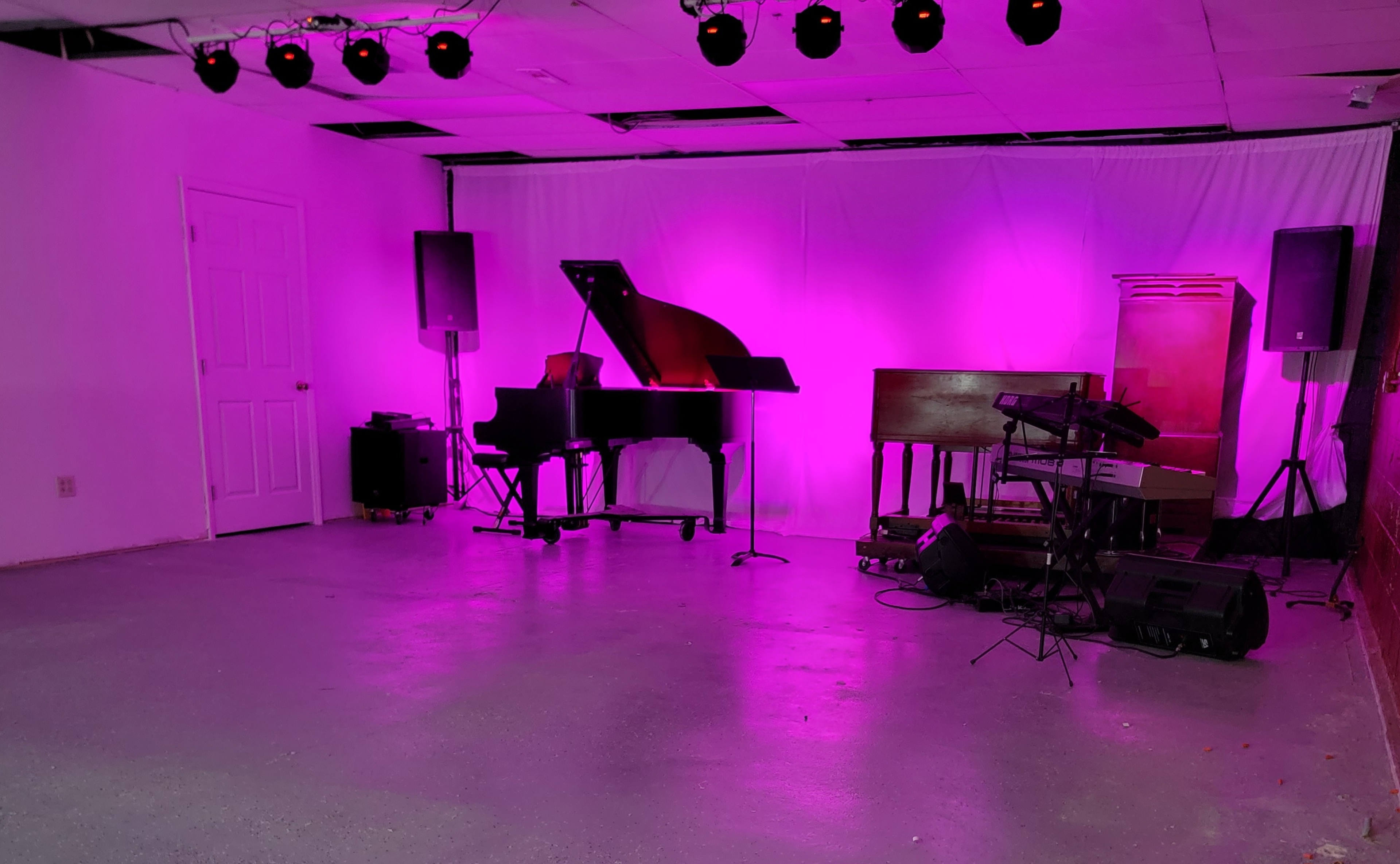 The image shows a small performance space with a grand piano, an organ, and various speakers, all illuminated by pink lighting.