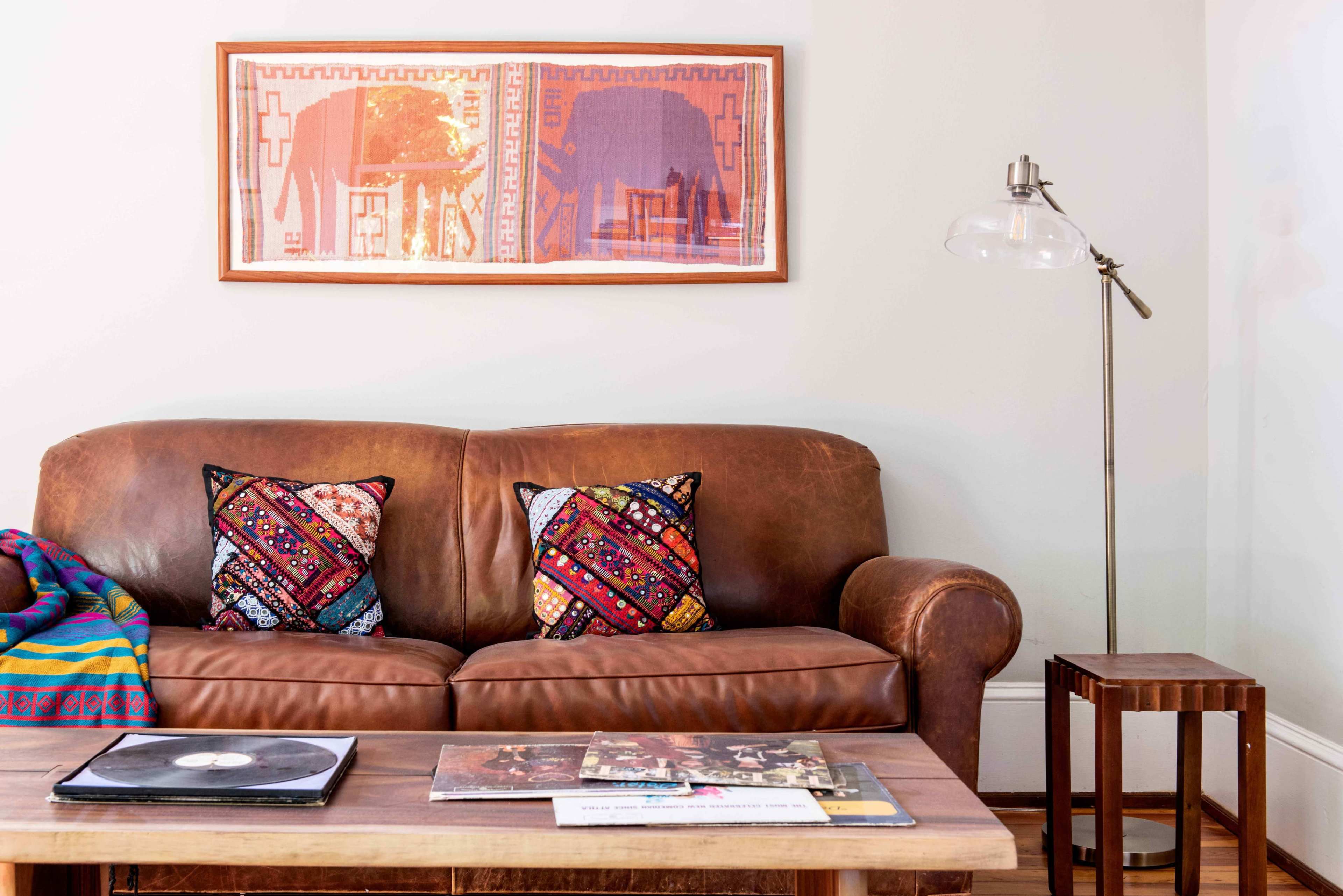 A brown leather sofa with patterned cushions is positioned beside a wooden coffee table stacked with magazines, under a framed artwork of elephants on the wall.