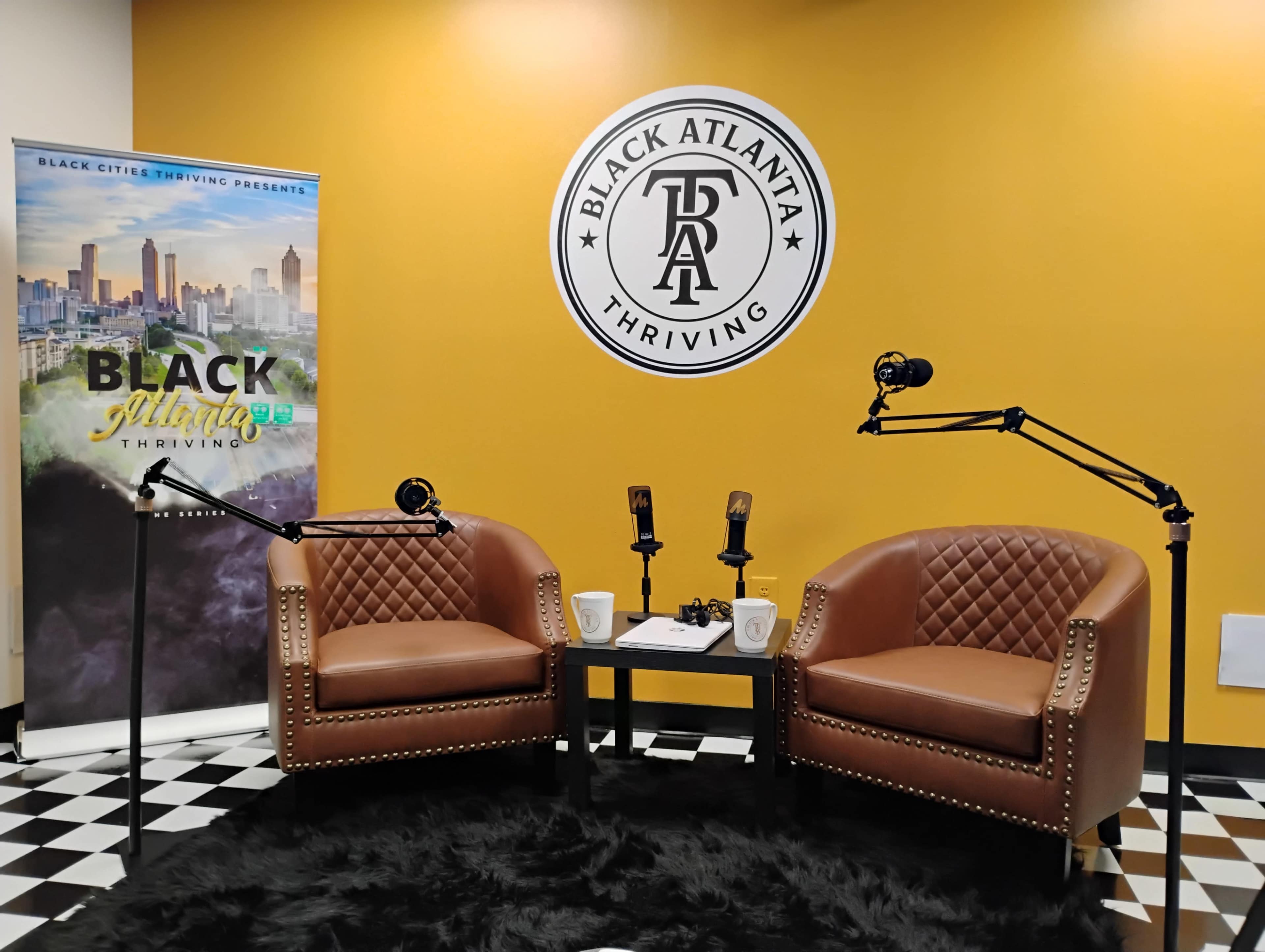 The image shows a cozy podcast studio with two brown leather chairs, two microphone stands, and a backdrop featuring a yellow wall with the logo "BLACK ATLANTA Thriving."