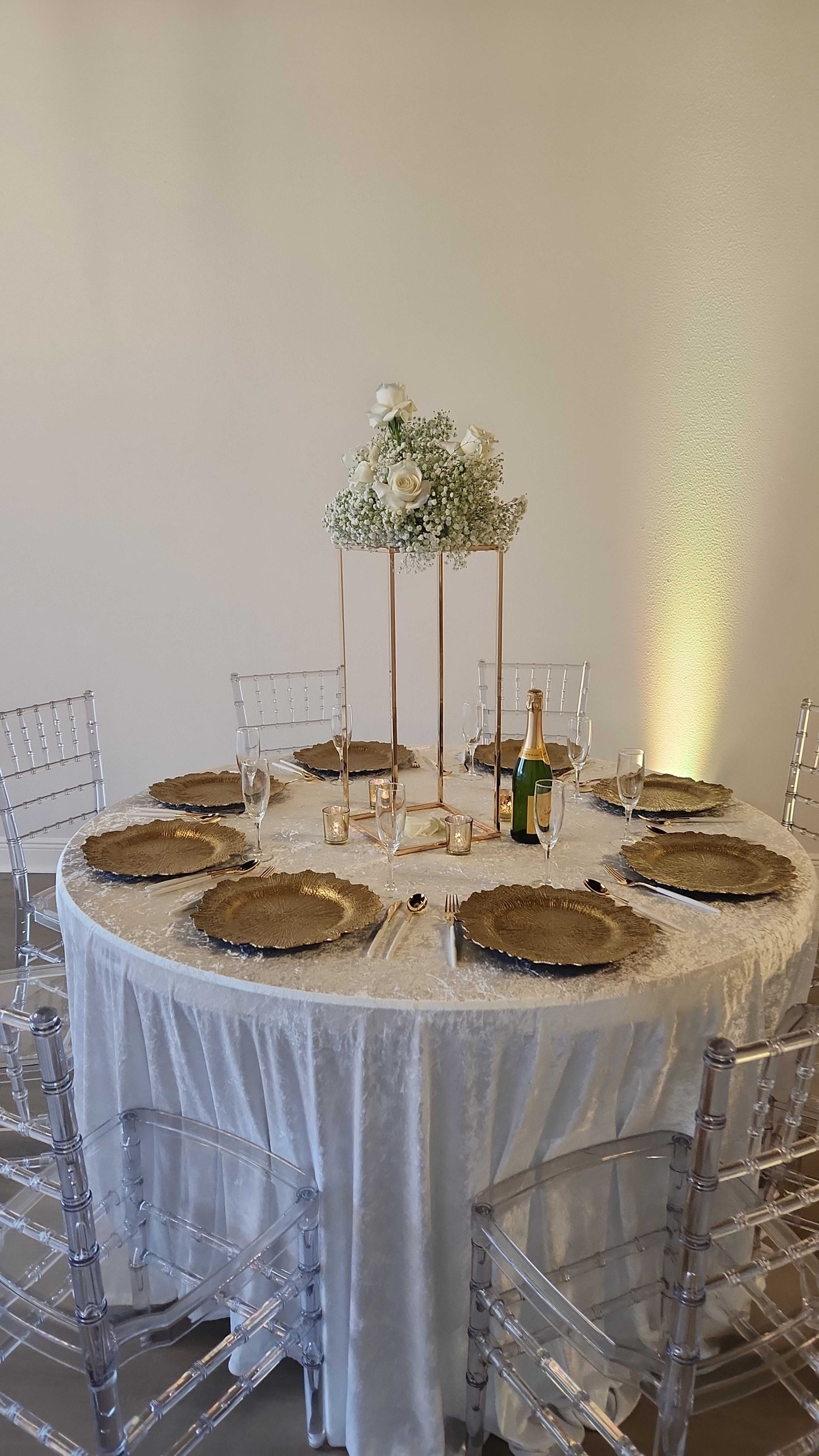 A round table is set with gold chargers, glassware, and a floral centerpiece, with a bottle of champagne nearby.