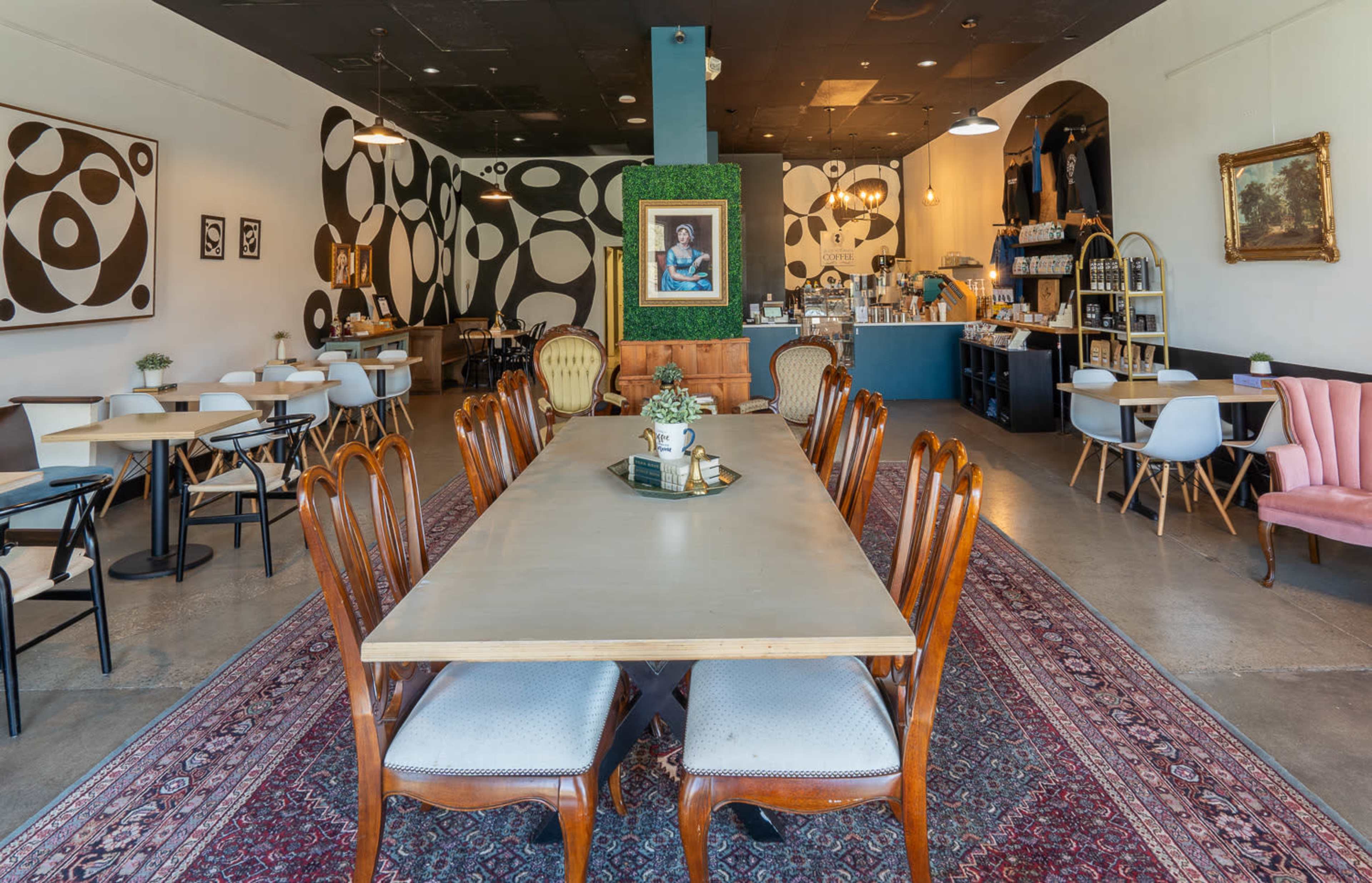 A contemporary café interior with a long wooden table, mixed seating arrangements, and patterned walls featuring abstract designs.
