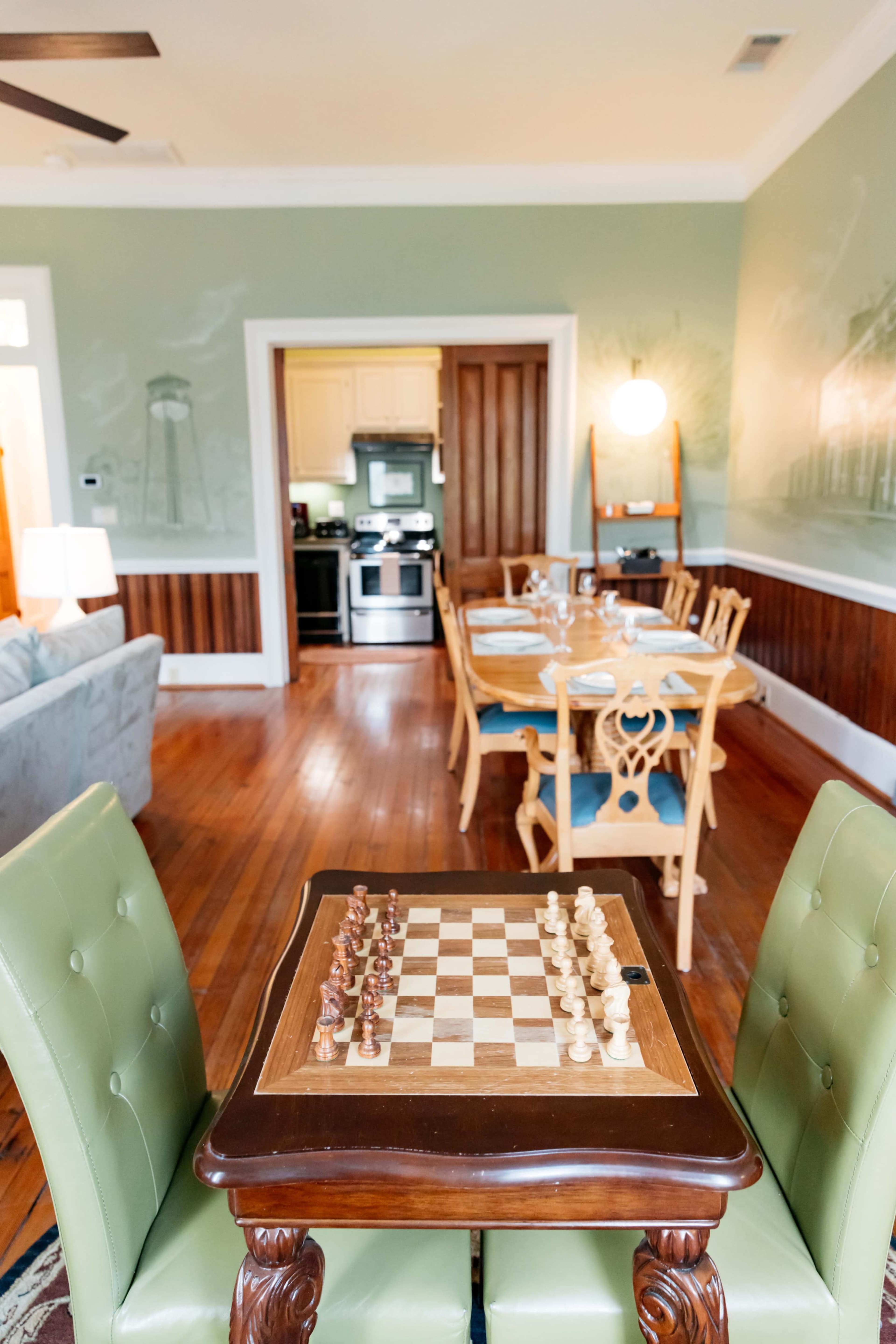 A chessboard sits on a wooden table in the foreground, with a dining area and kitchen visible in the background.