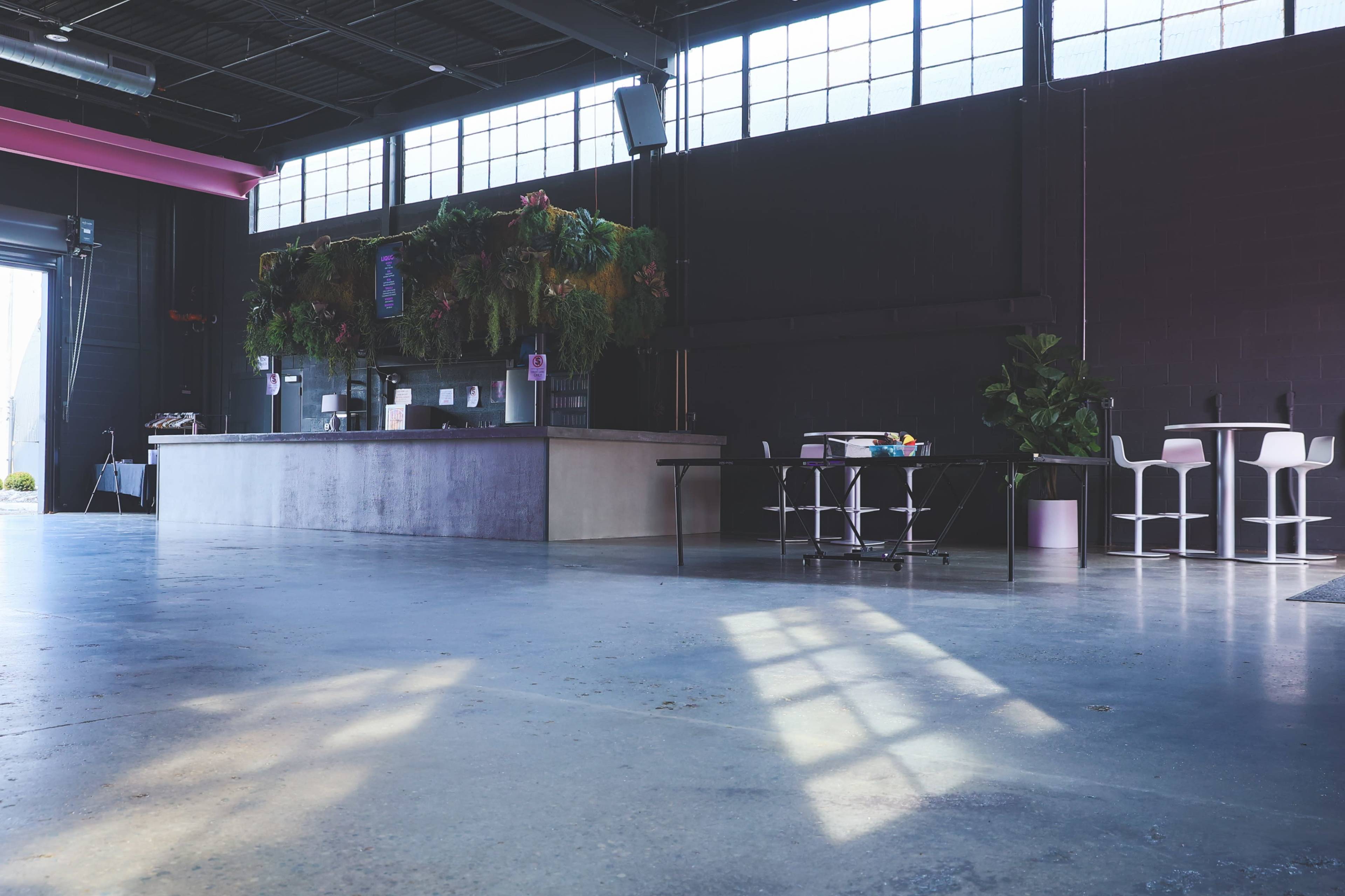 A spacious interior with a bar area featuring a lush vertical garden, surrounded by tables and chairs under large windows allowing natural light to enter.