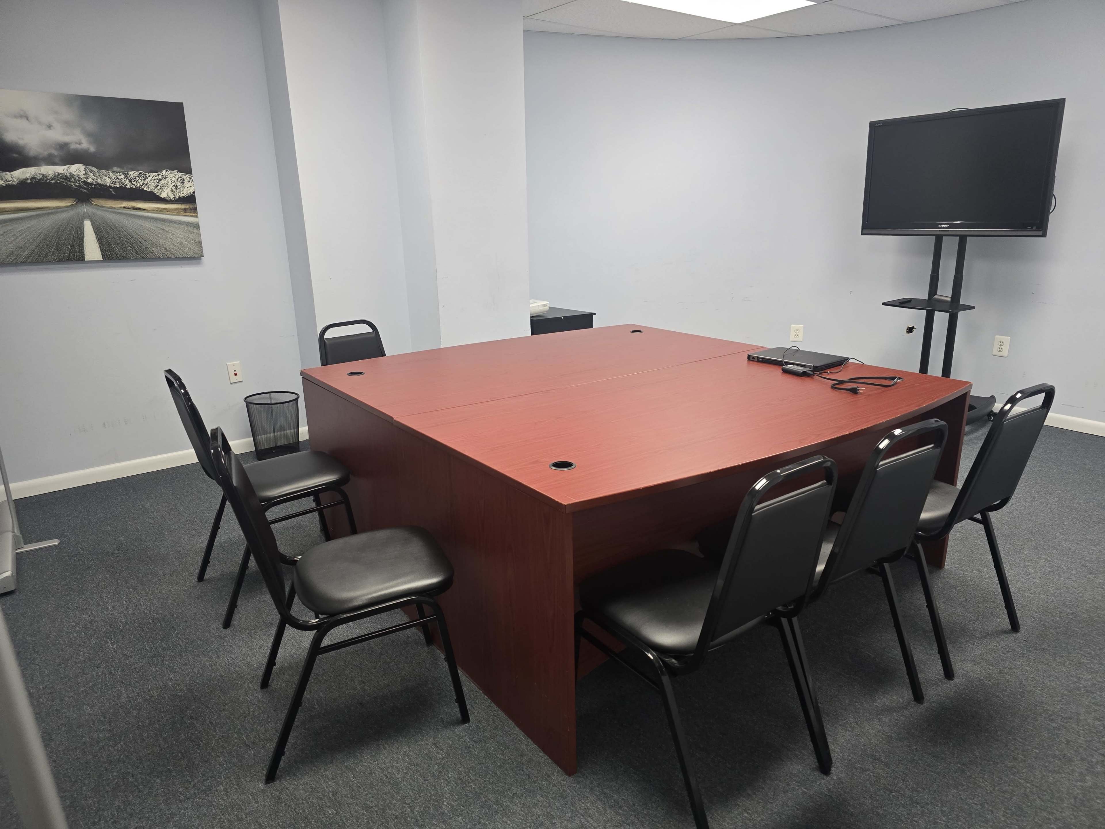 A rectangular conference table with six black chairs is positioned in a sparsely furnished meeting room, featuring a wall-mounted television and a landscape photograph.