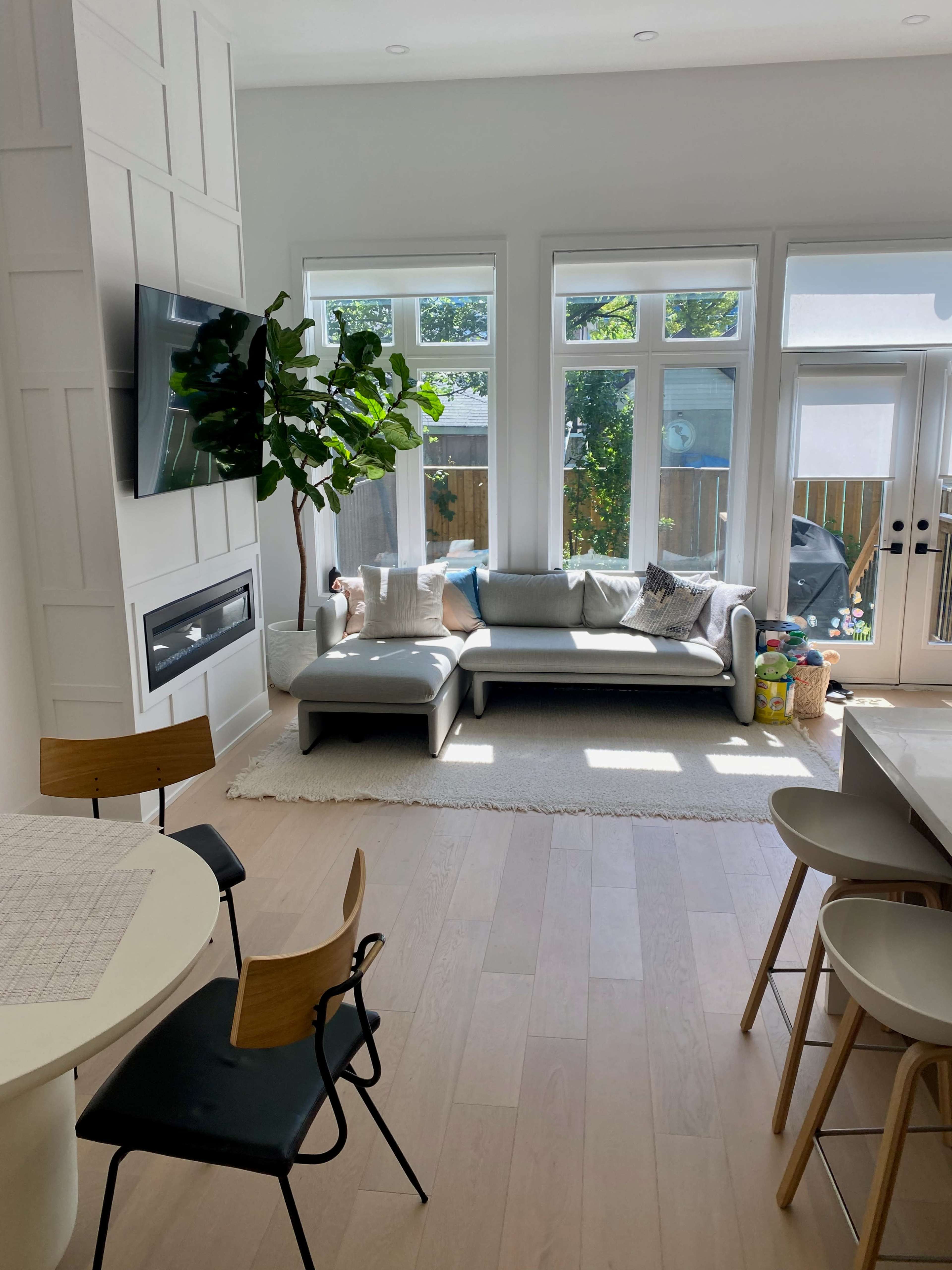 A modern living room with a gray sofa, large windows allowing natural light, and a small dining area with a round table and chairs.