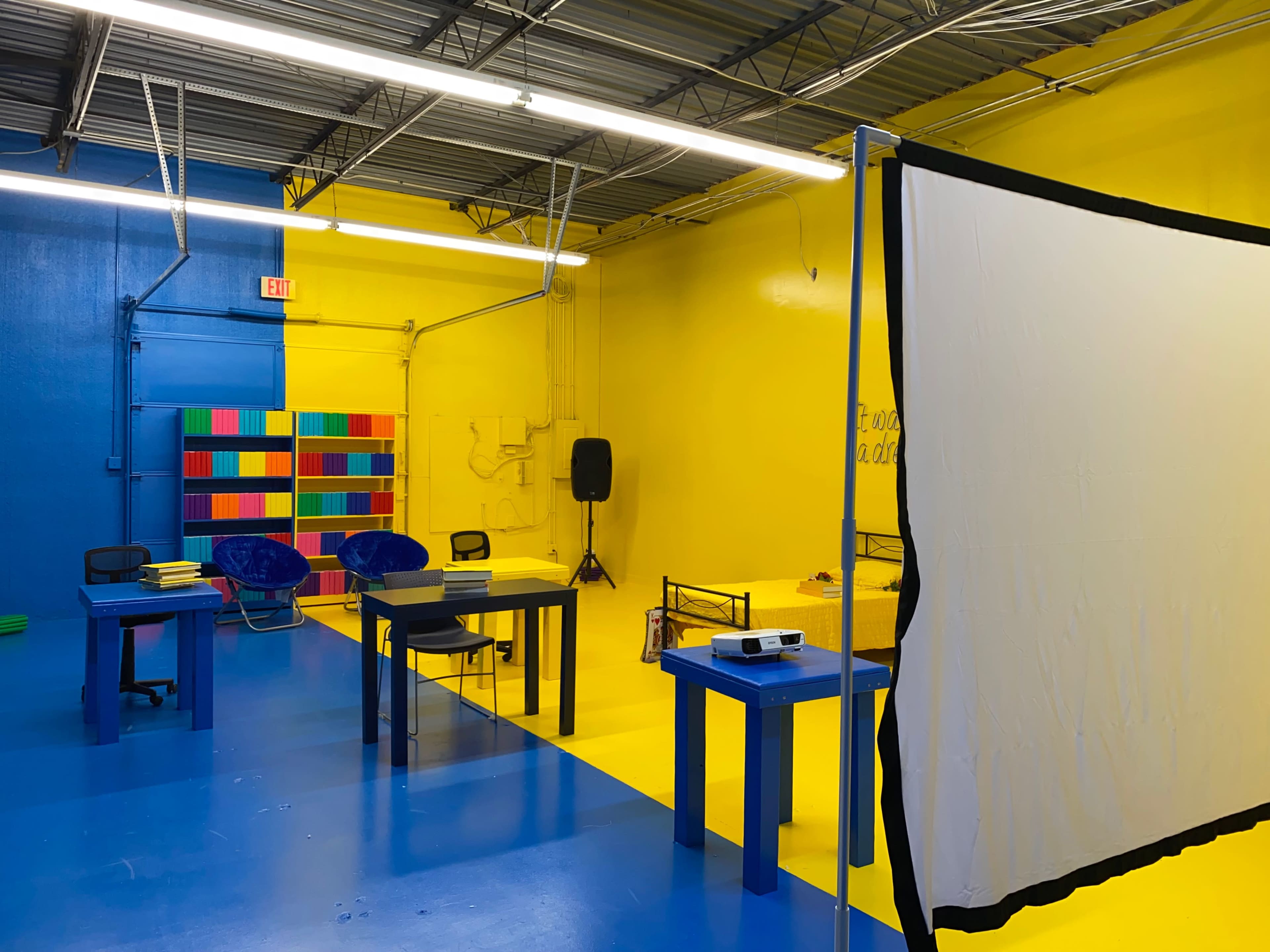 A brightly colored room with yellow and blue walls, featuring several tables, chairs, and a projector screen on a stand.