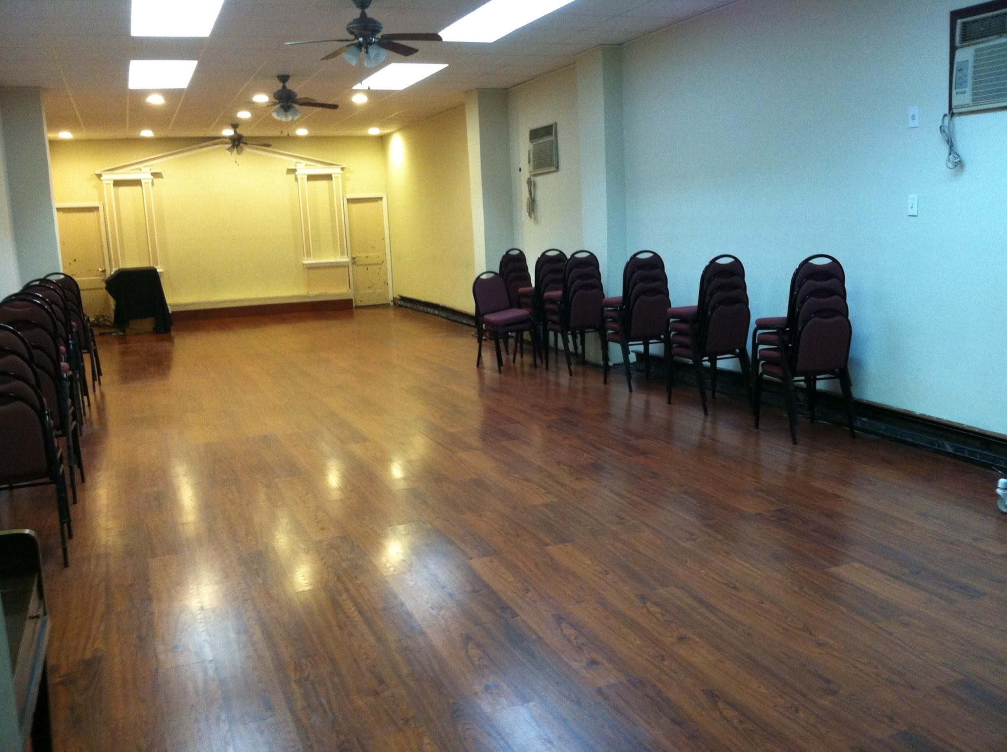 A spacious room with a polished wooden floor and rows of stacked chairs along the walls.