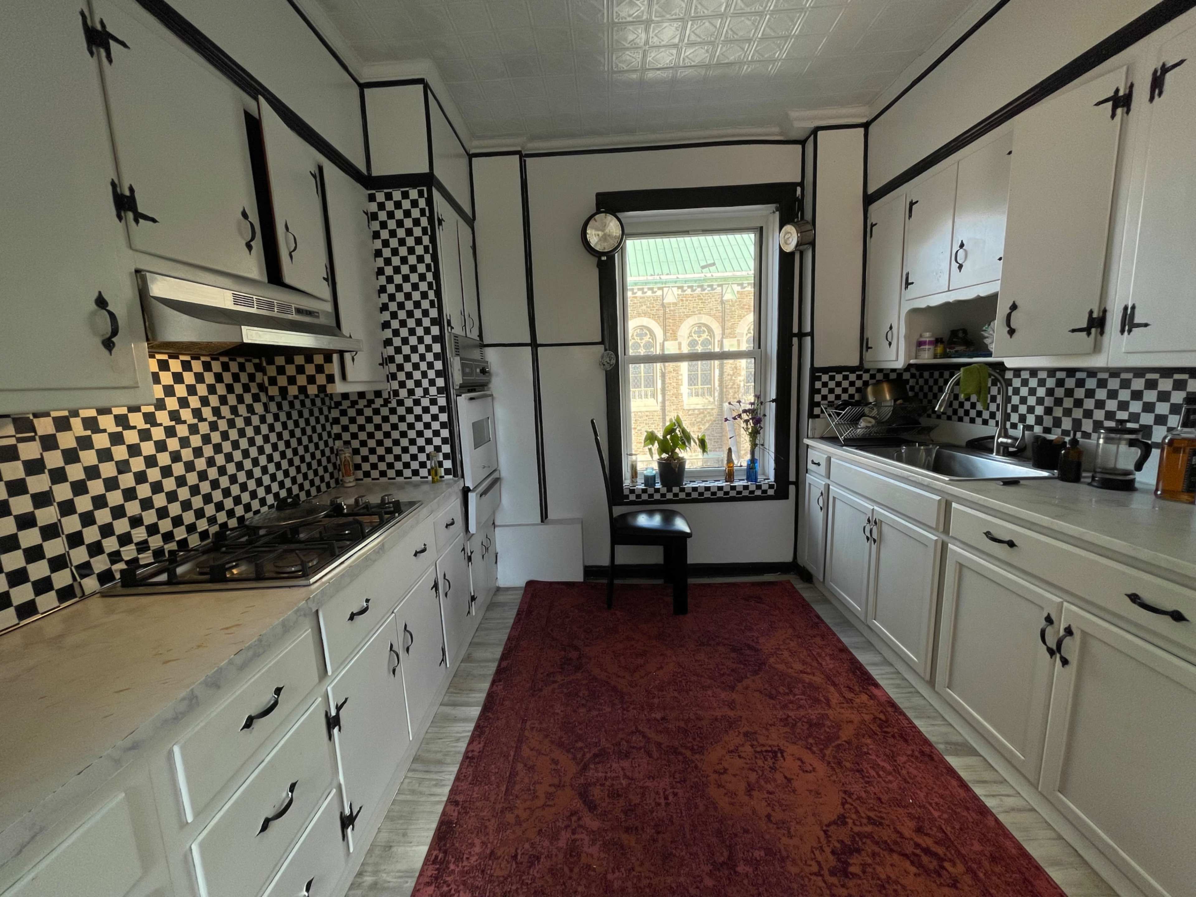 A kitchen is shown with black and white checkered walls, a gas stove, and a window overlooking an exterior view, featuring cabinets and a rug on the floor.