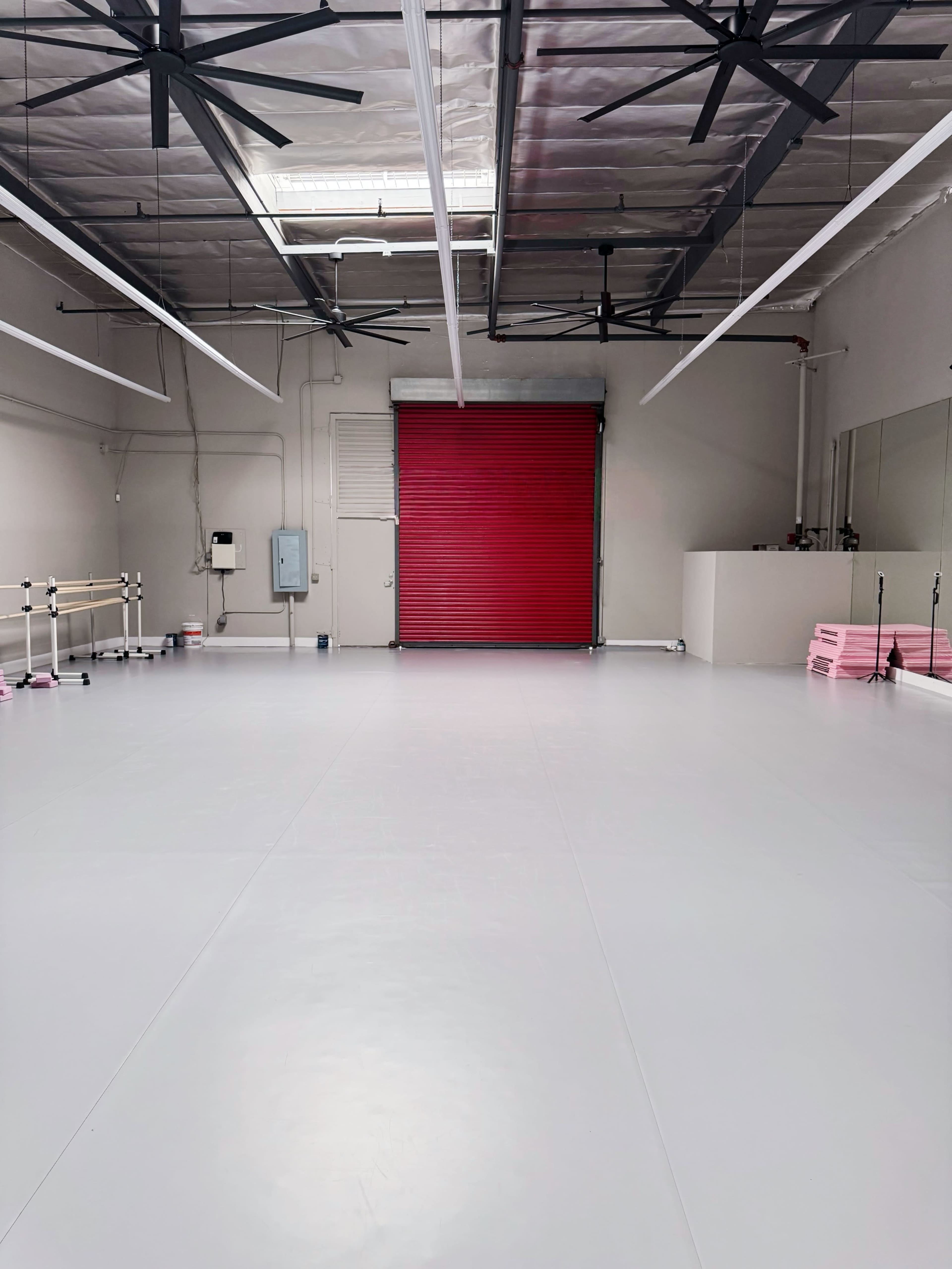 The image shows a spacious, empty studio with a gray floor, large ceiling fans, and a red roll-up door in the back.