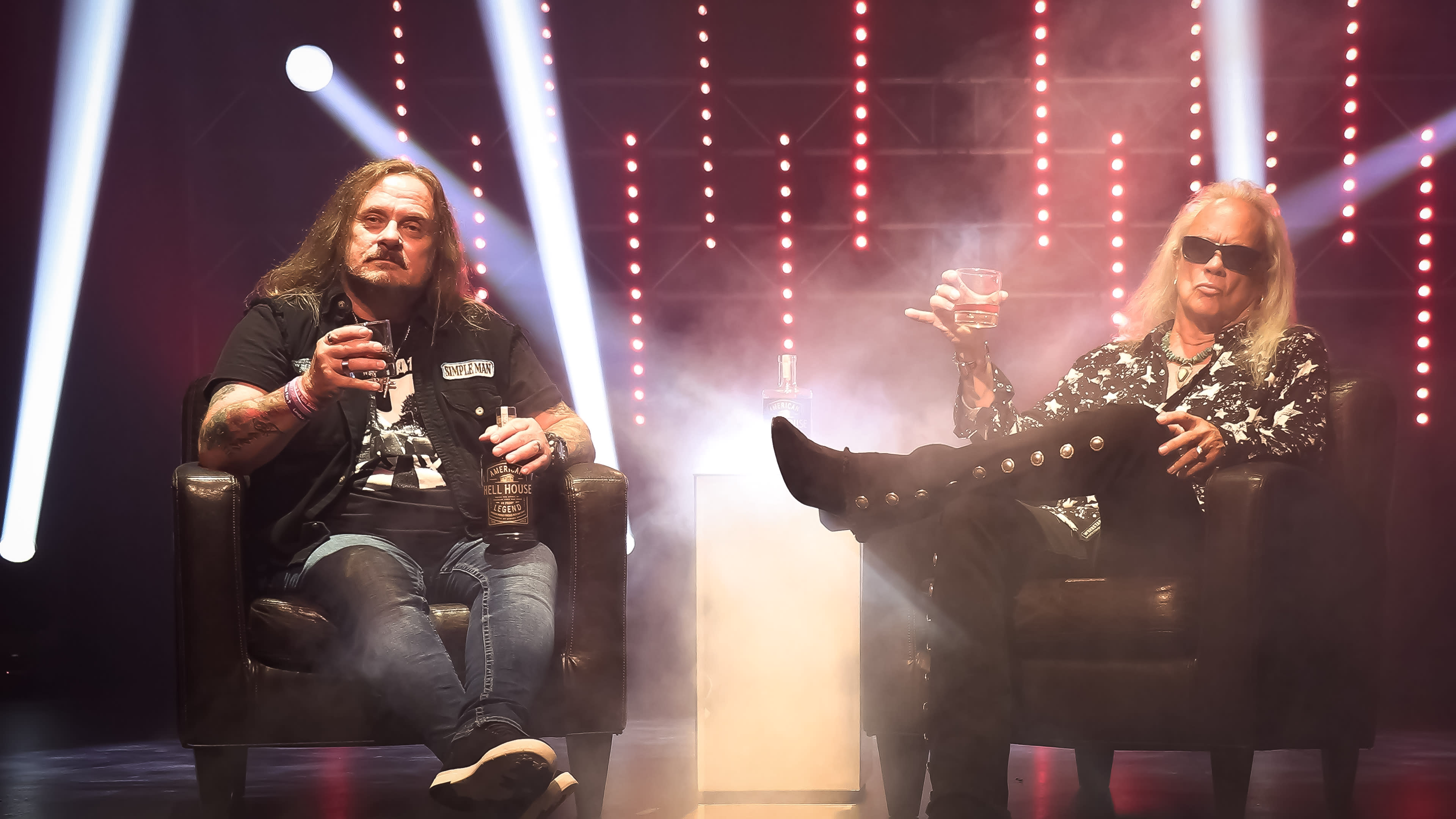Two men sit in armchairs, each holding a drink, amidst a backdrop of stage lights and fog.