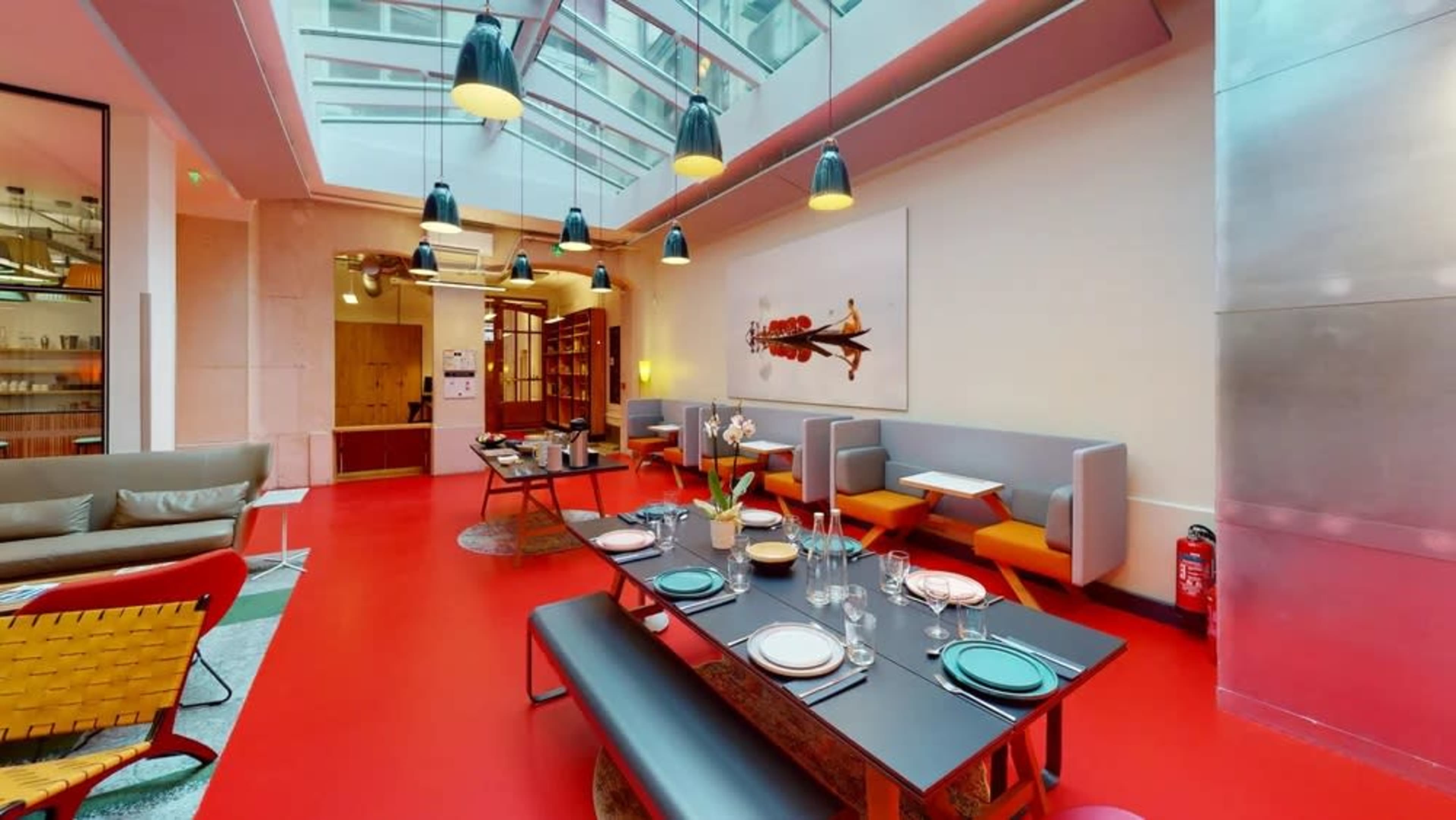 A modern dining area with a red floor, a long table set for a meal, and surrounding seating against a backdrop of large windows.