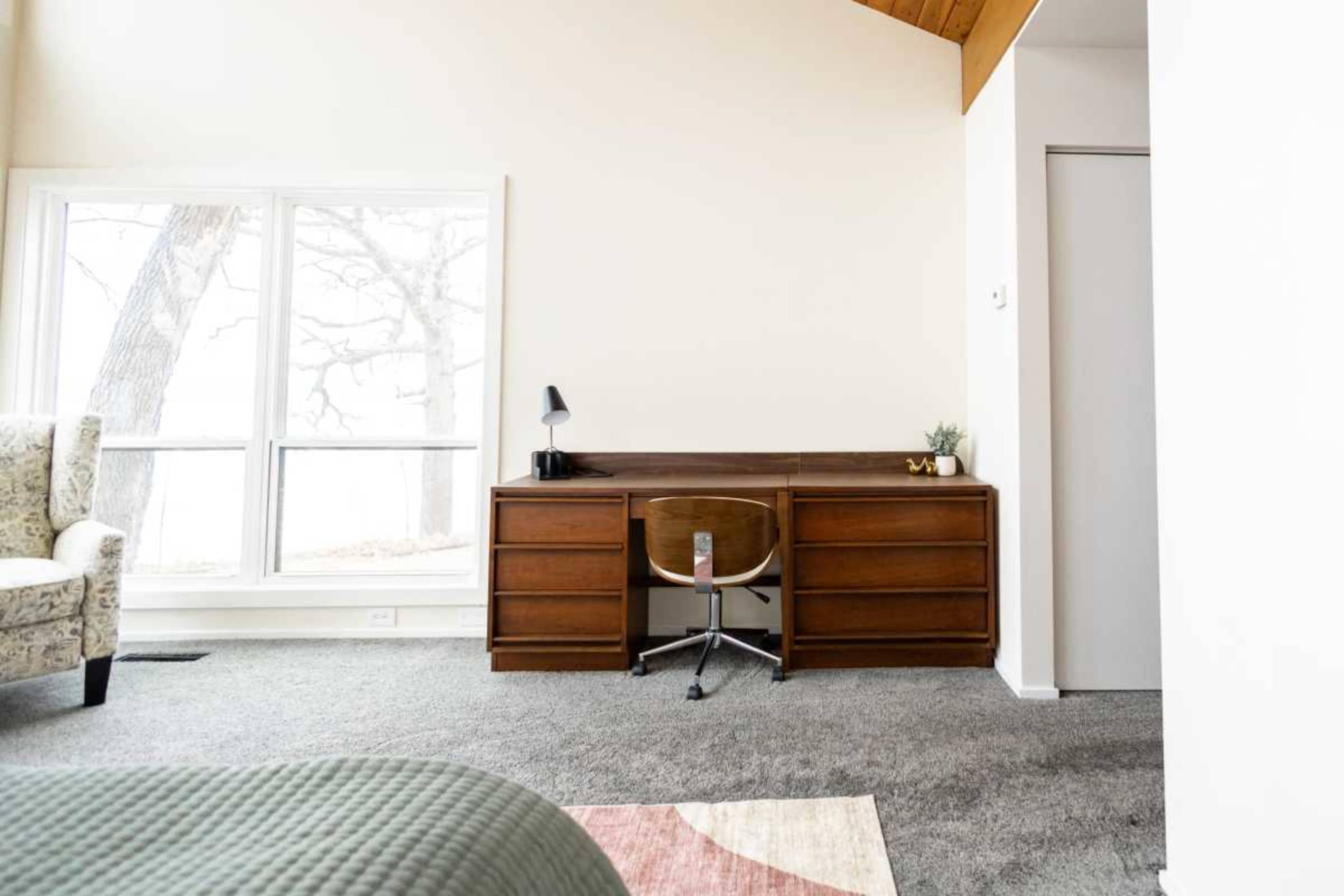 The image shows a minimalist bedroom featuring a wooden desk with a chair, a lamp, and a small plant beside a large window.