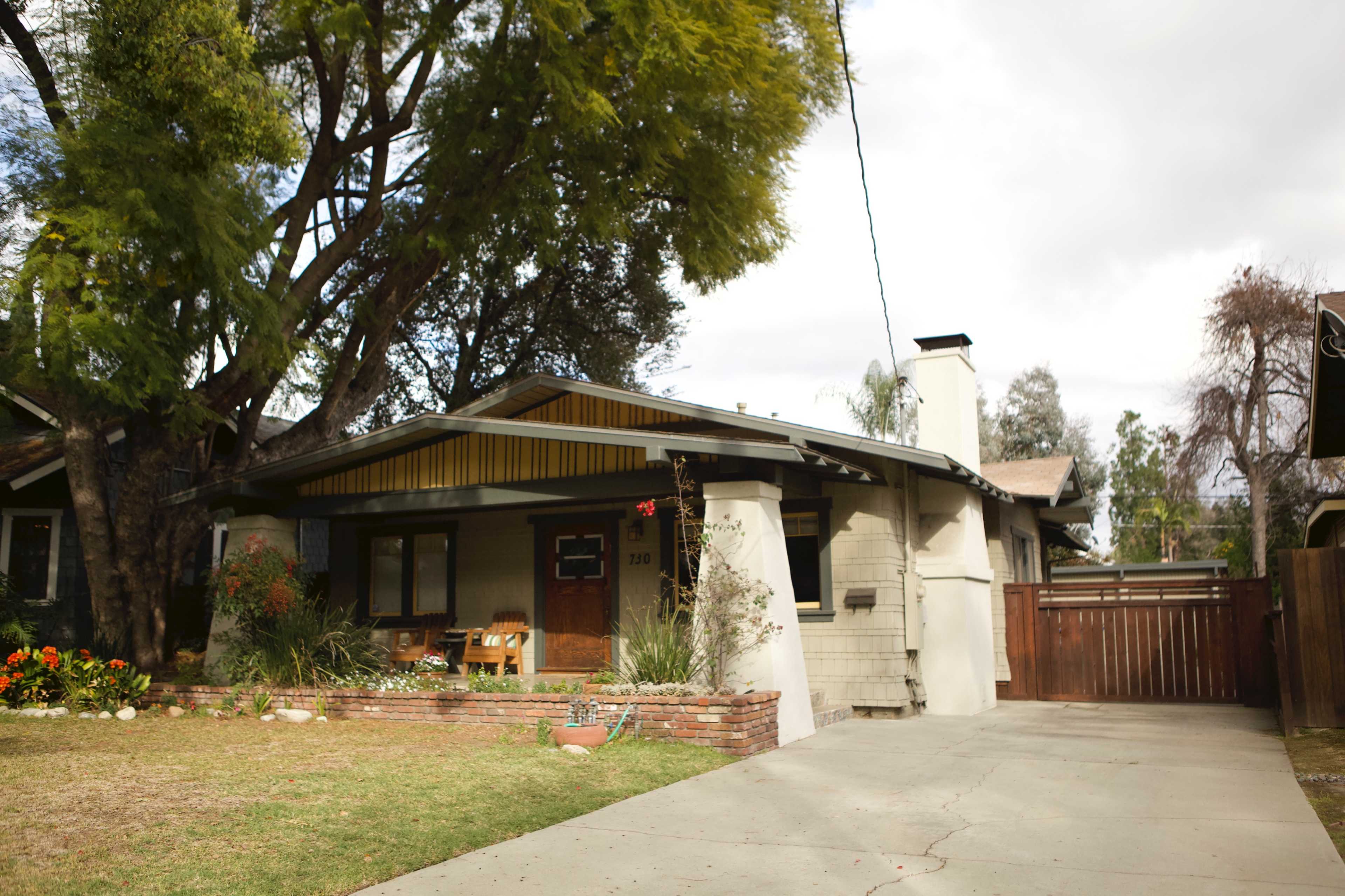 A single-story house with a front garden, a wooden porch, and a driveway leads to a gate.