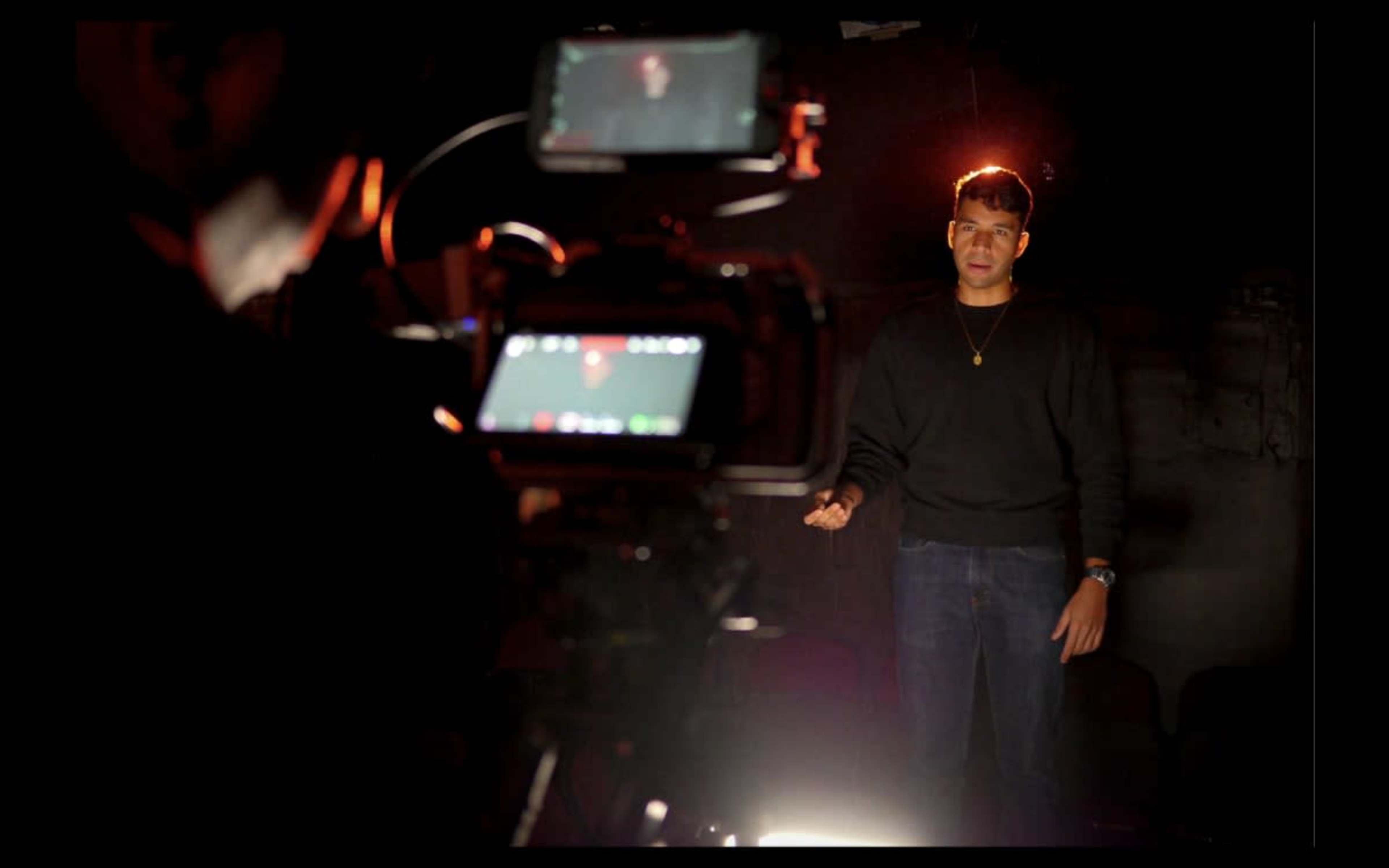 A man stands in front of a camera, speaking under soft lighting in a dimly lit room.