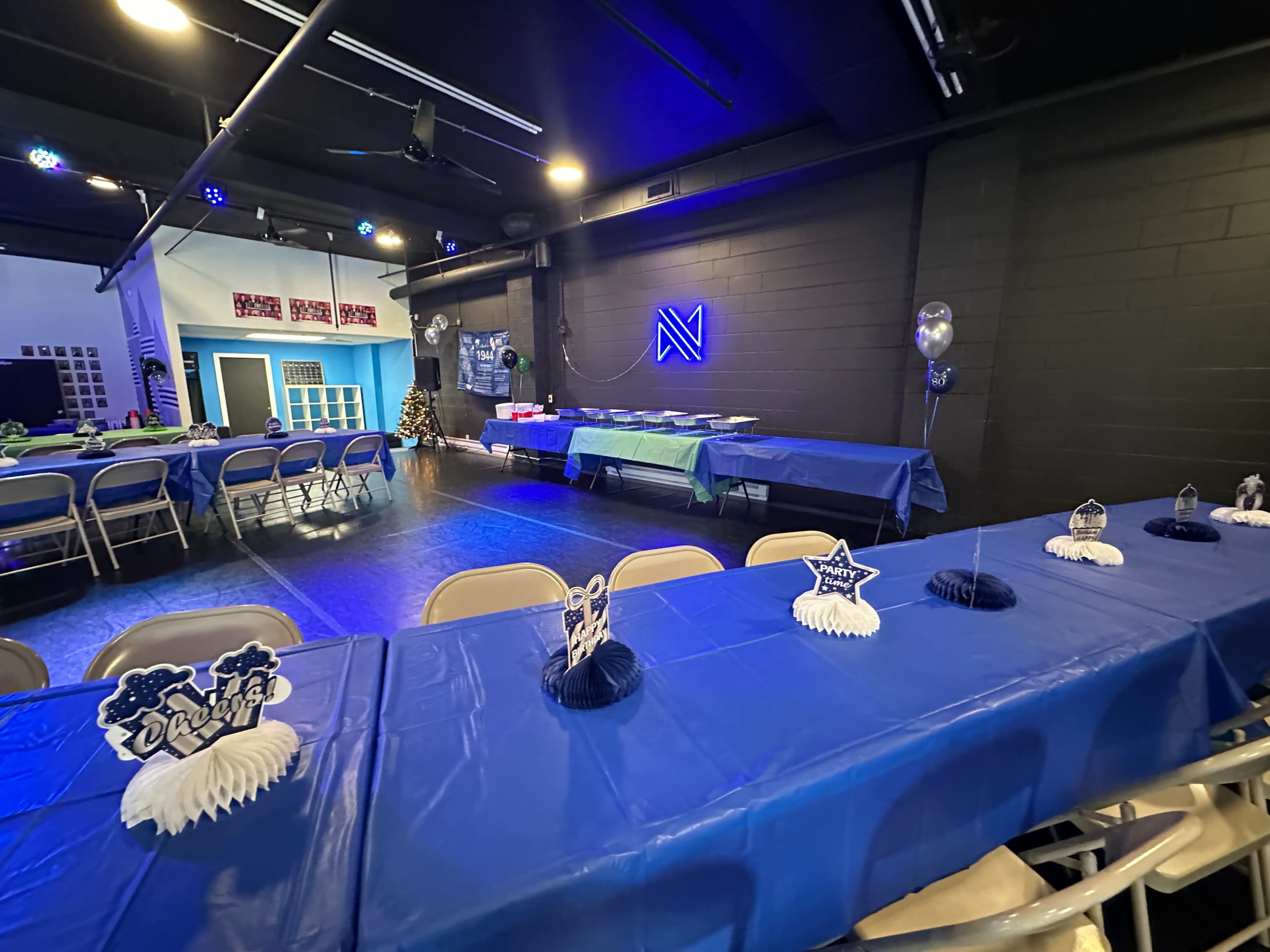 The image shows a decorated event space with blue tables set up for a gathering, featuring centerpieces and a buffet table in the background.