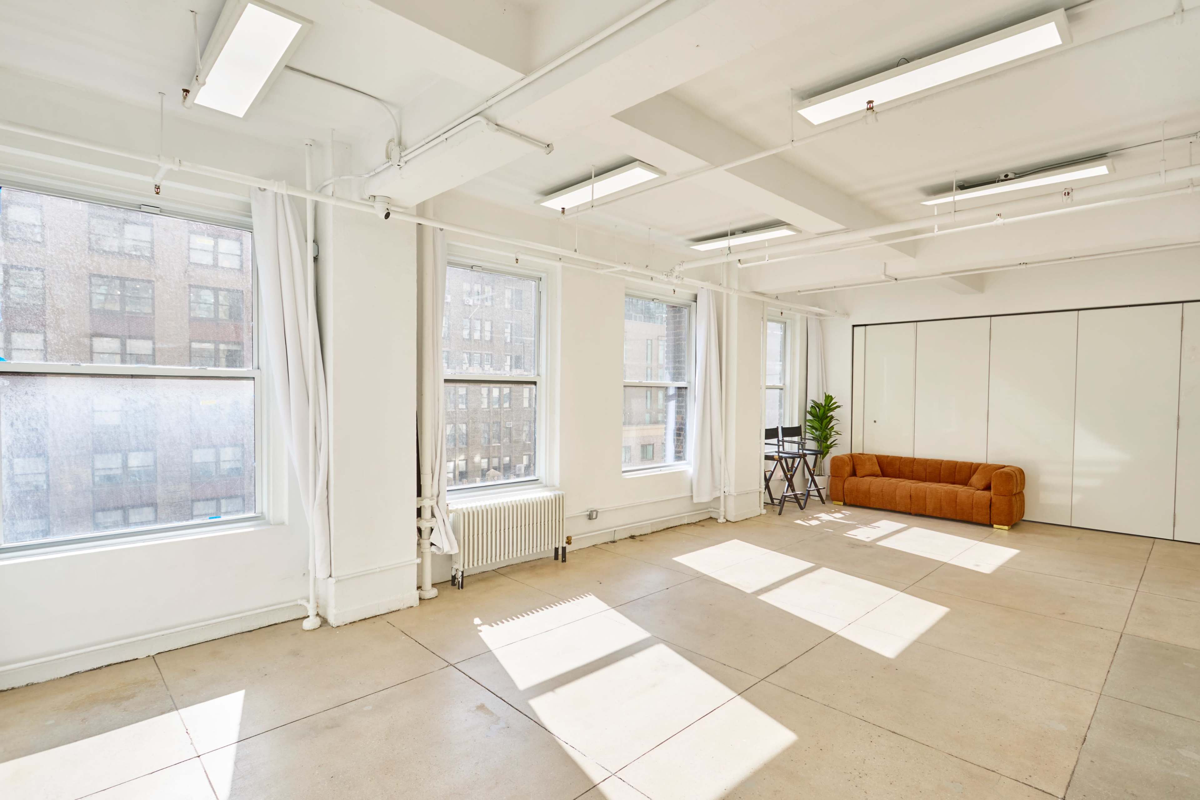 The image shows a bright, empty room with large windows, white walls, a concrete floor, and a brown sofa along one side.